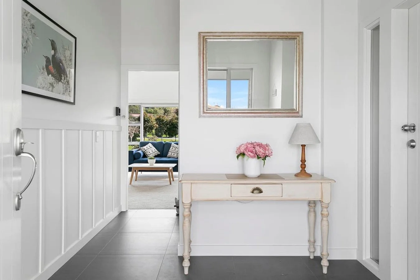 View of an entryway in a home with a beige console table, a vase of pink flowers, a mirror, a lamp, and a door on the right. Through the doorway, a living room with a blue sofa, pillows, and a window showing a porch and trees outside.