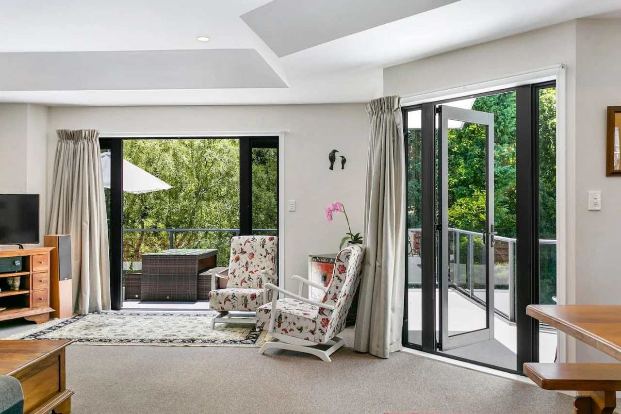 Living room with sliding glass doors leading to a balcony, two floral armchairs, a wooden cabinet with a TV, a potted orchid, and outdoor green trees.