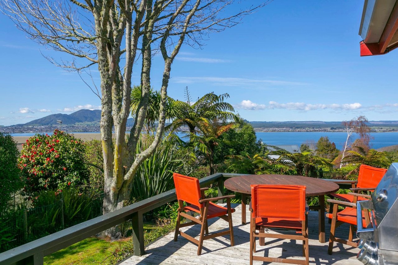 A wooden deck with orange chairs around a round table overlooks a scenic view of trees, a river, and mountains in the distance on a sunny day.