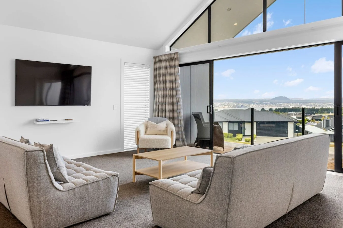 Living room with large sliding glass doors opening to a balcony with a scenic view, two beige sofas, a white armchair, a wooden coffee table, and a mounted flat-screen TV on a white wall.