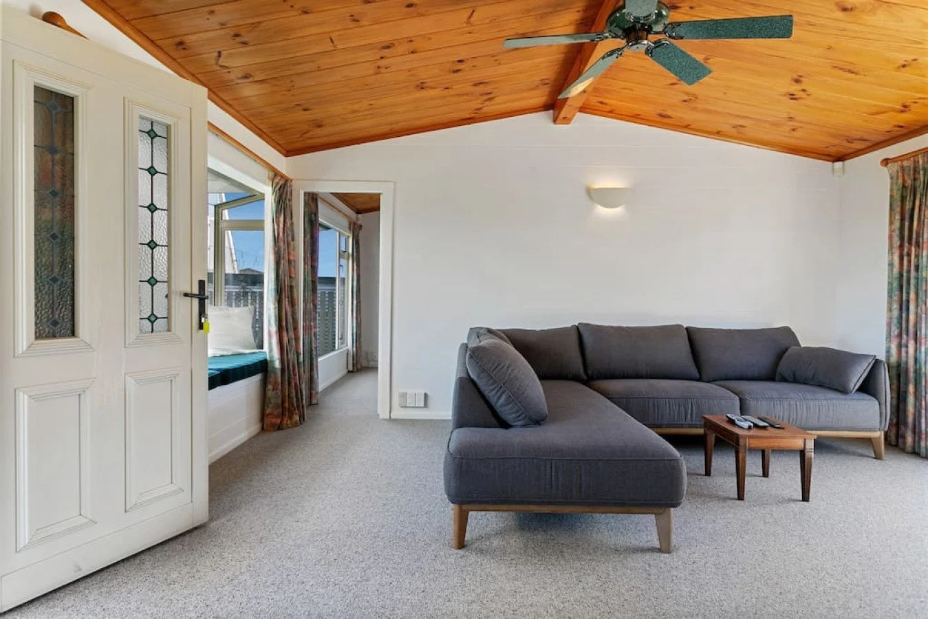Living room with a gray sectional sofa, wooden coffee table, ceiling fan, white walls, carpeted floor, and large windows with floral curtains.