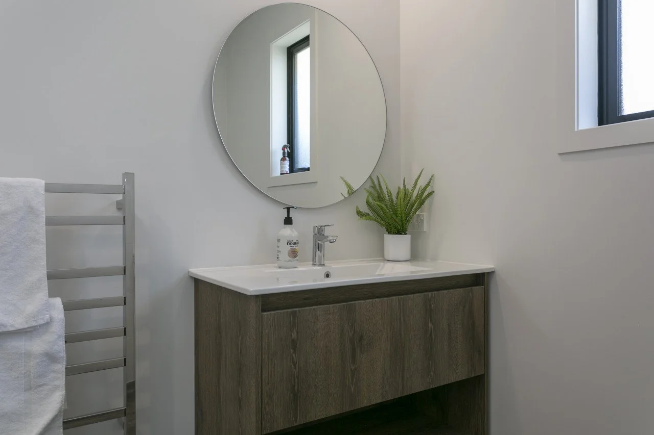 Modern bathroom with gray towel rack, white walls, and a round mirror above a wooden vanity with a white sink. There is a potted green plant on the vanity and a bottle of soap. Two small windows are visible, providing natural light.