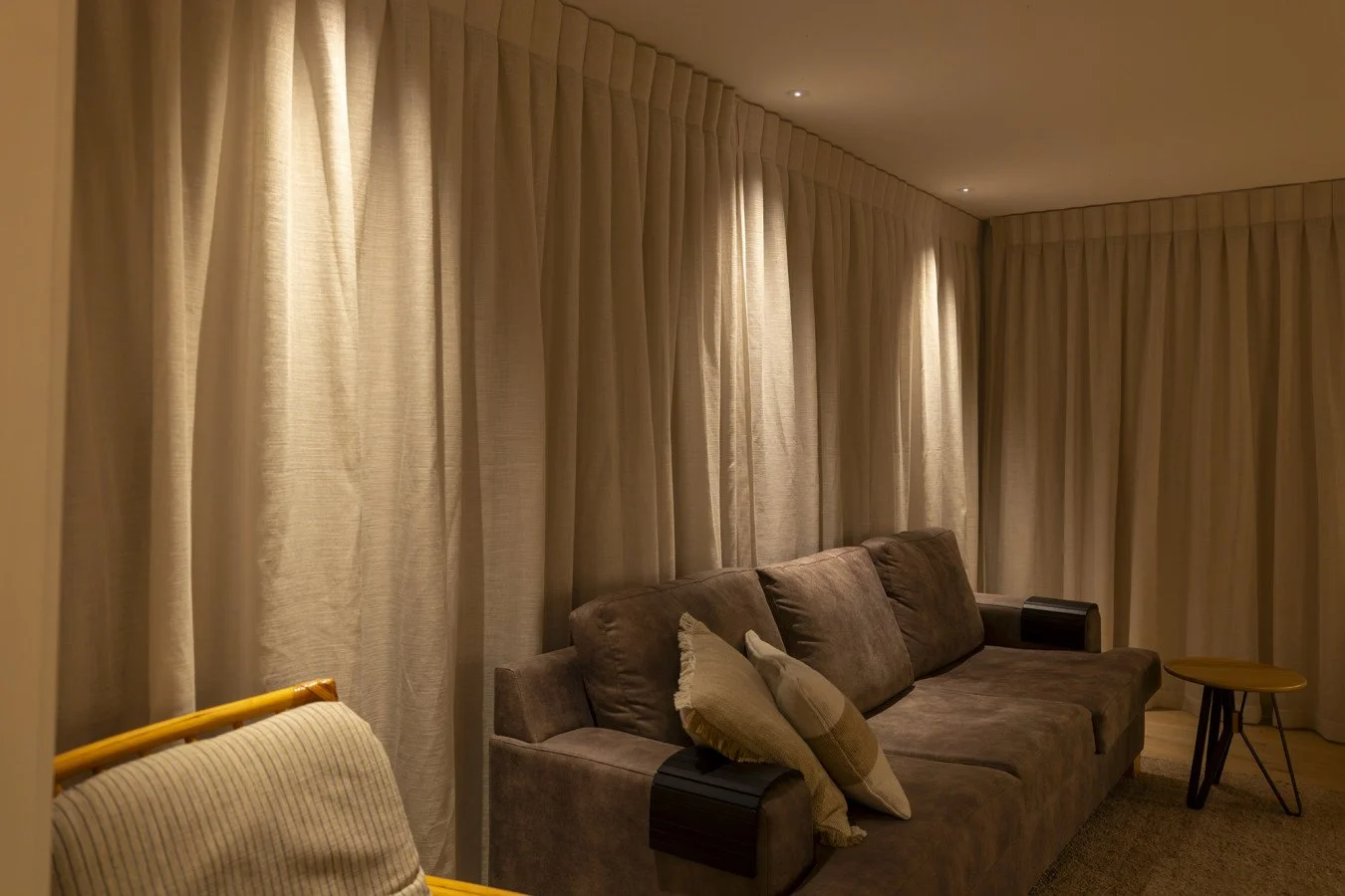 Living room with beige curtains, a brown sofa with pillows, and small side tables.
