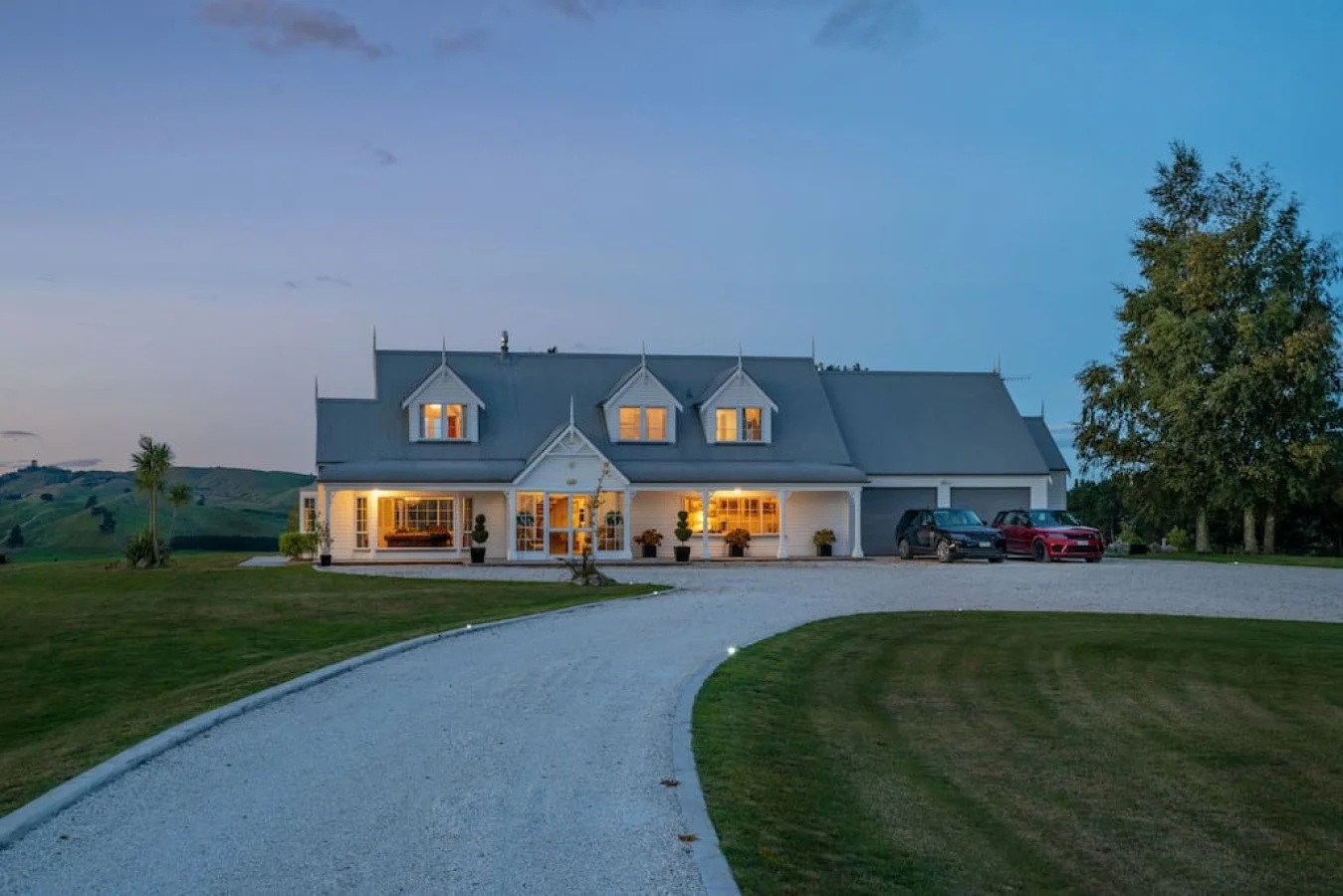 A large white house with a gray roof and multiple dormer windows, illuminated from inside, with a winding gravel driveway, green lawn, and trees at dusk.