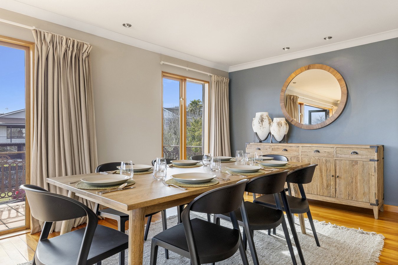 Dining room with a wooden table set for six, black chairs, a light wood sideboard, decorative vases, a round mirror, two large windows with beige curtains, hardwood flooring, blue accent wall, and ceiling lights.