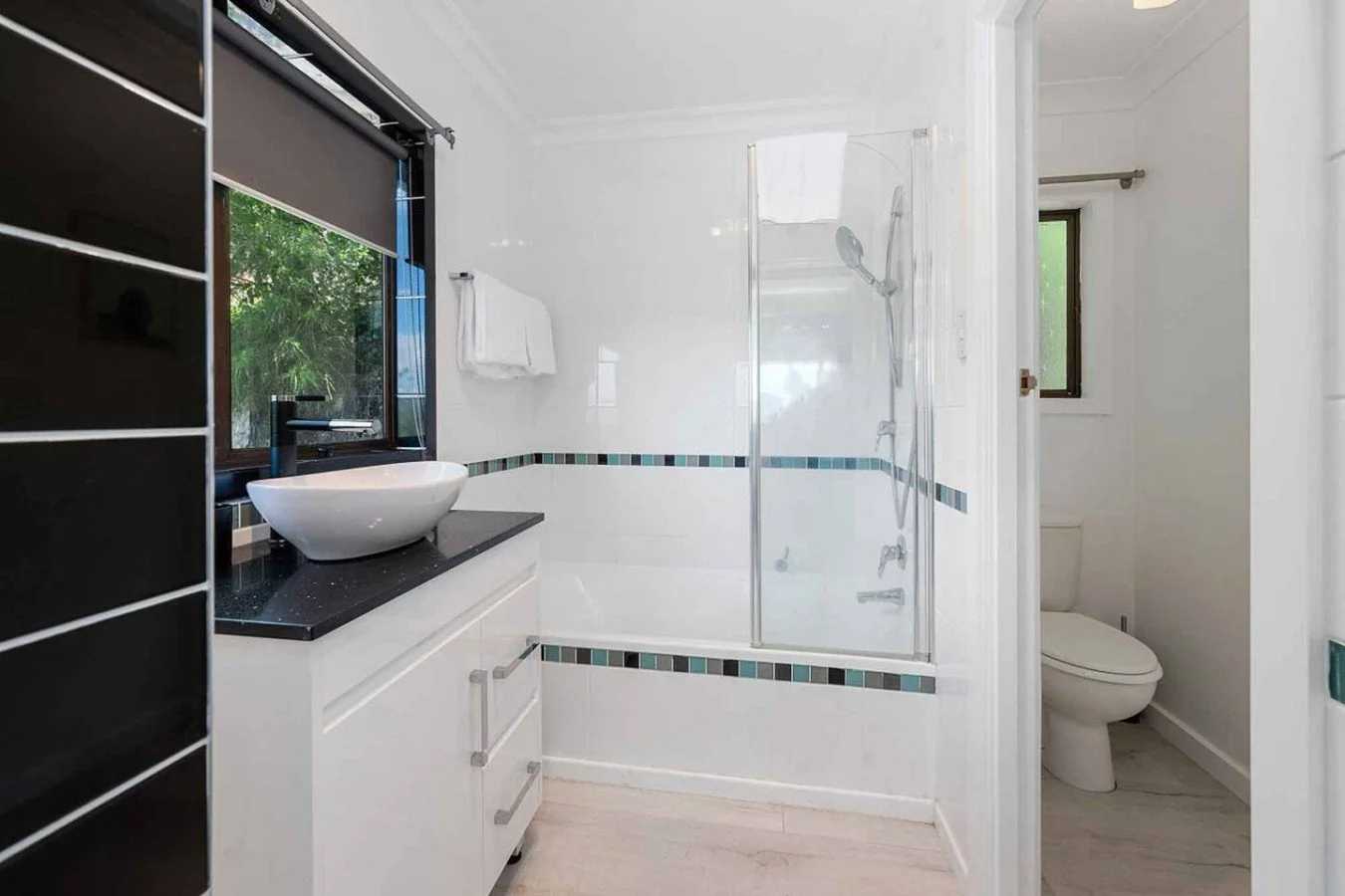Modern bathroom with white walls, a black countertop with a vessel sink, a glass-enclosed shower, a window, and a toilet.