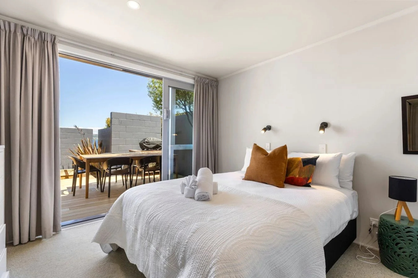 A bedroom with a large bed, white bedding, and accent pillows, opens to a balcony with a table and chairs, and outdoor decor.