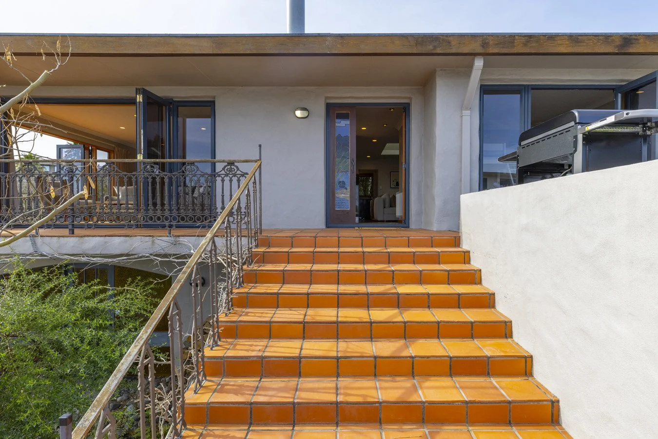 Exterior view of a house entrance with orange tiled stairs, glass door, balcony with iron railing, and a barbecue grill on the right.
