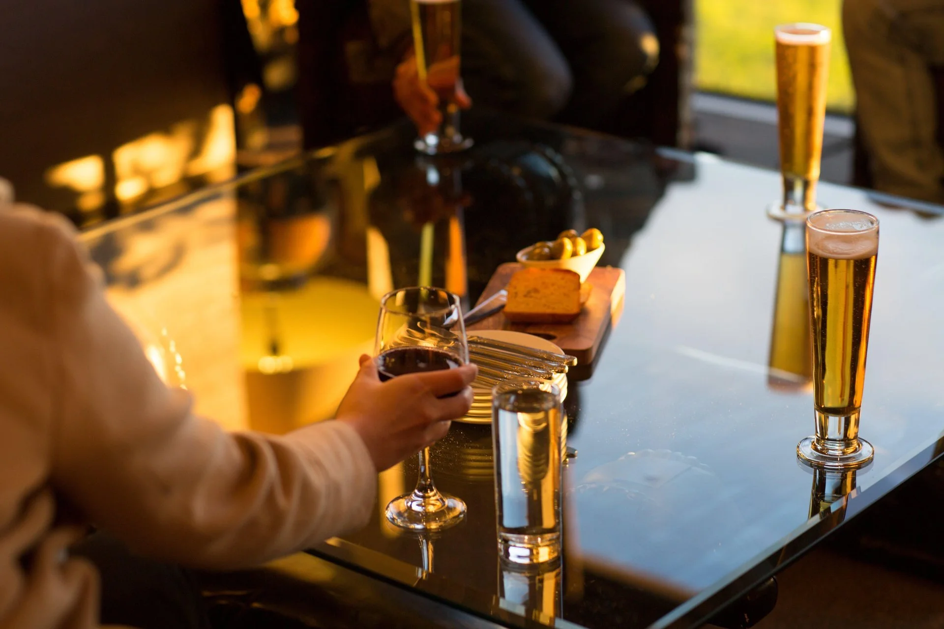 A person holding a glass of red wine sitting at a reflective glass table. On the table are three tall glasses of beer, a small plate with green olives, a block of cheese, and some breadsticks. The setting appears warm with sunlight coming through a window.