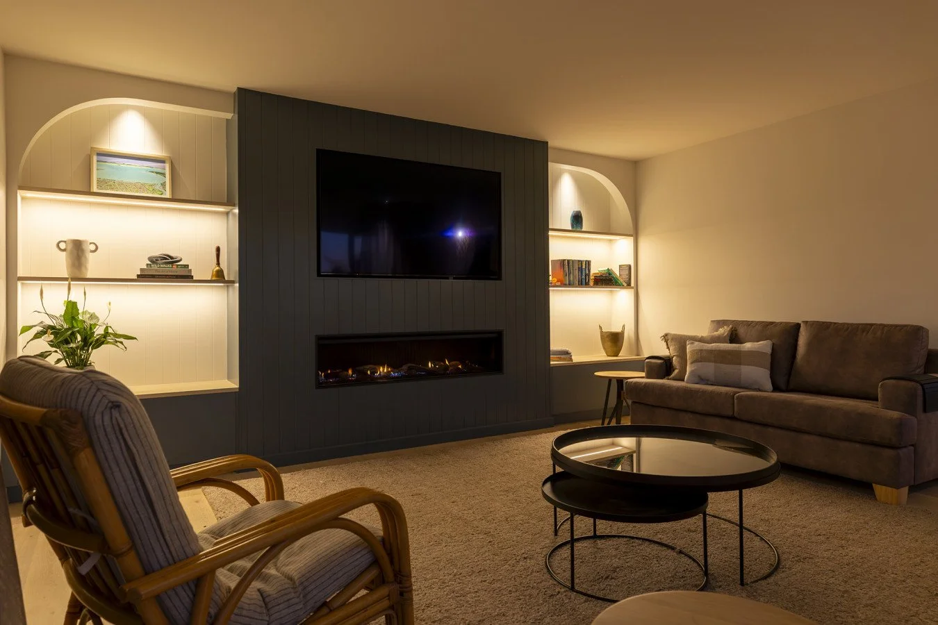 Living room with a gray couch, a rattan armchair, a round black coffee table, built-in shelves with books and decor, and a mounted flat-screen TV above a fireplace. Warm lighting, beige walls, and carpeted floor.