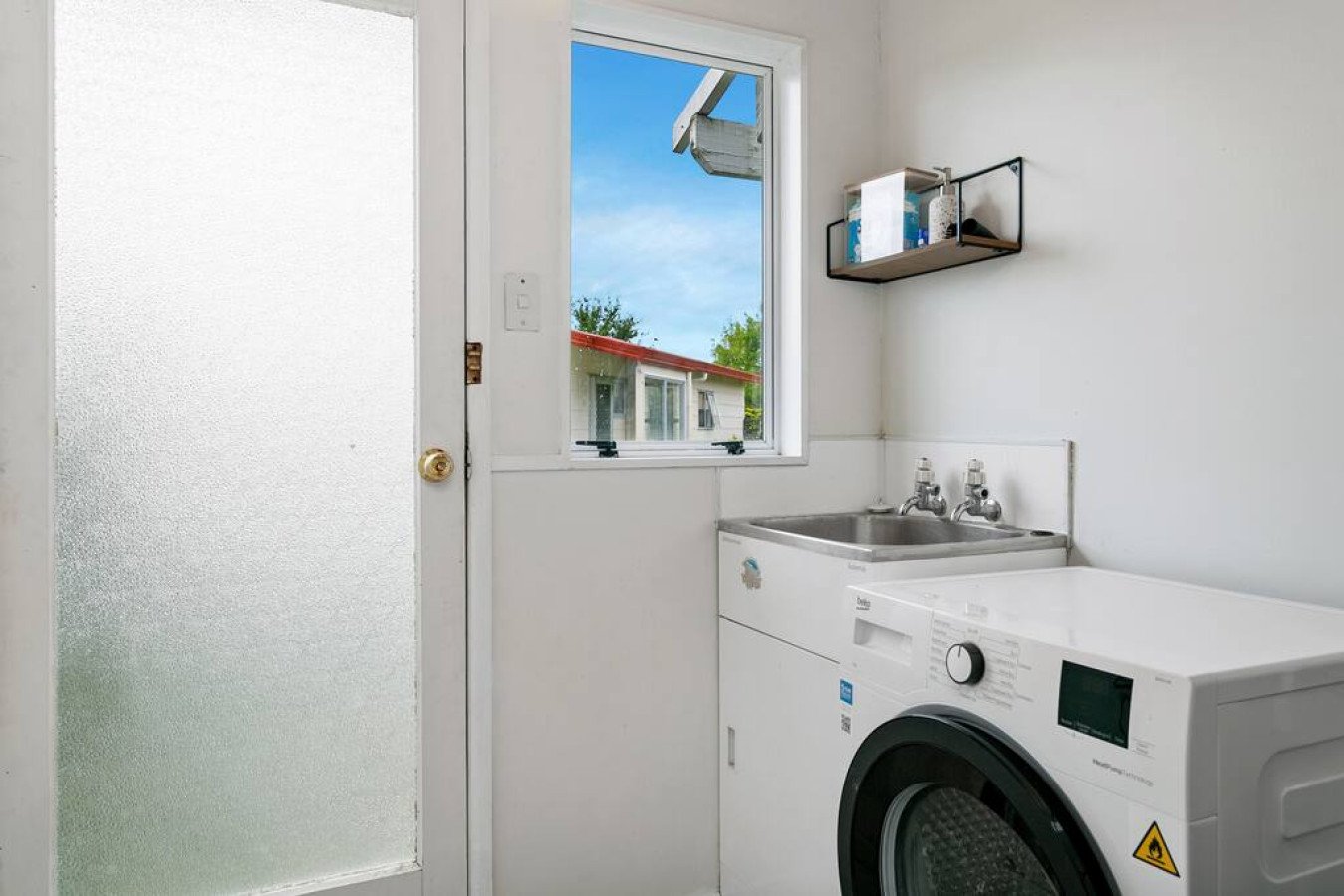 Laundry room with washing machine, utility sink, and frosted door, window showing outdoor view, wall-mounted shelf with supplies.