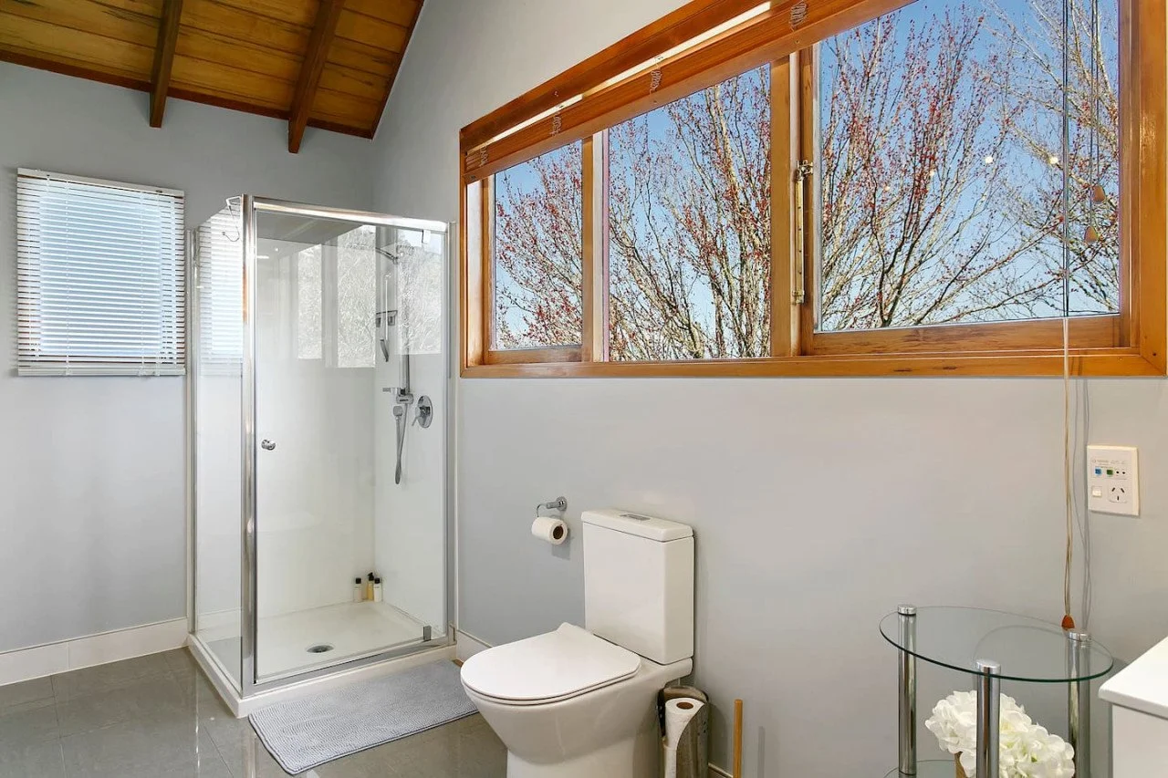 Bathroom with a glass shower enclosure, a white toilet, a window with a view of trees outside, and a small glass table with white flowers.