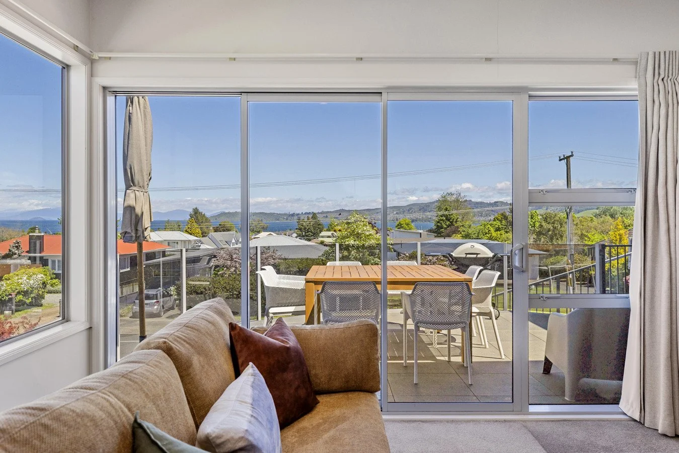 Living room with large sliding glass door leading to a balcony. The balcony has outdoor furniture, a barbecue grill, and scenic views of trees, houses, and mountains in the background.