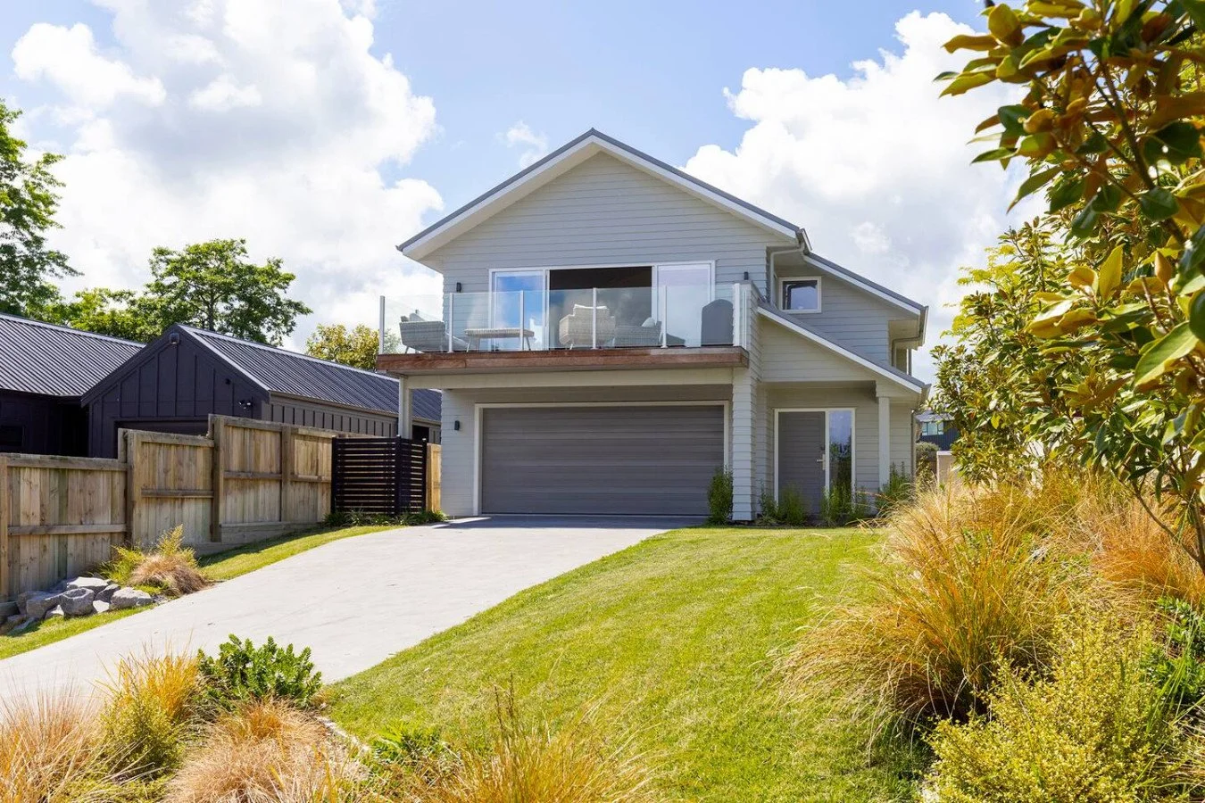 A modern two-story house with a gray garage door, a balcony with glass railing, and a well-maintained lawn and garden in front.