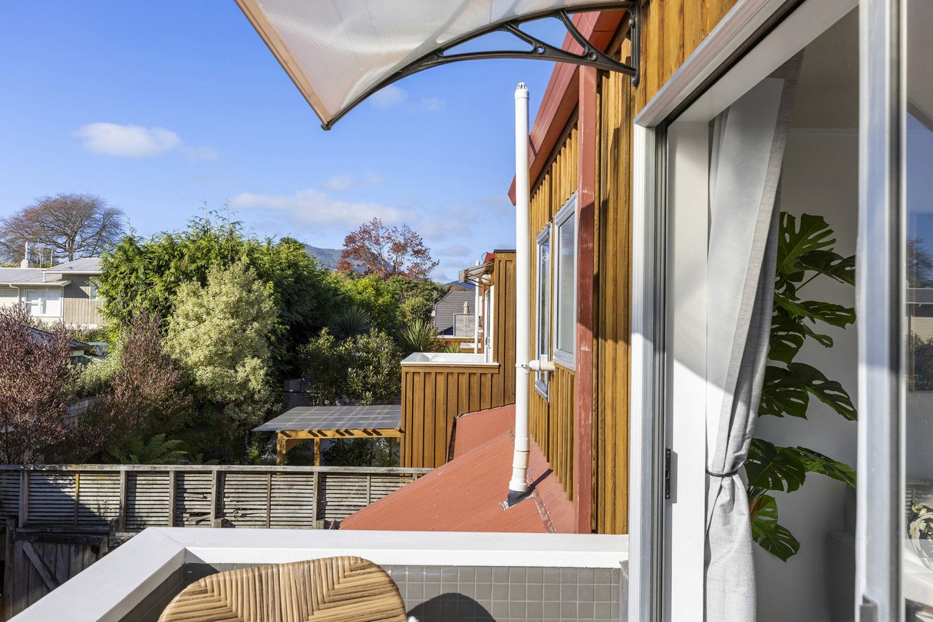 View from a balcony showing a wooden house exterior with large sliding glass door, an indoor plant, and trees in the background under a blue sky.