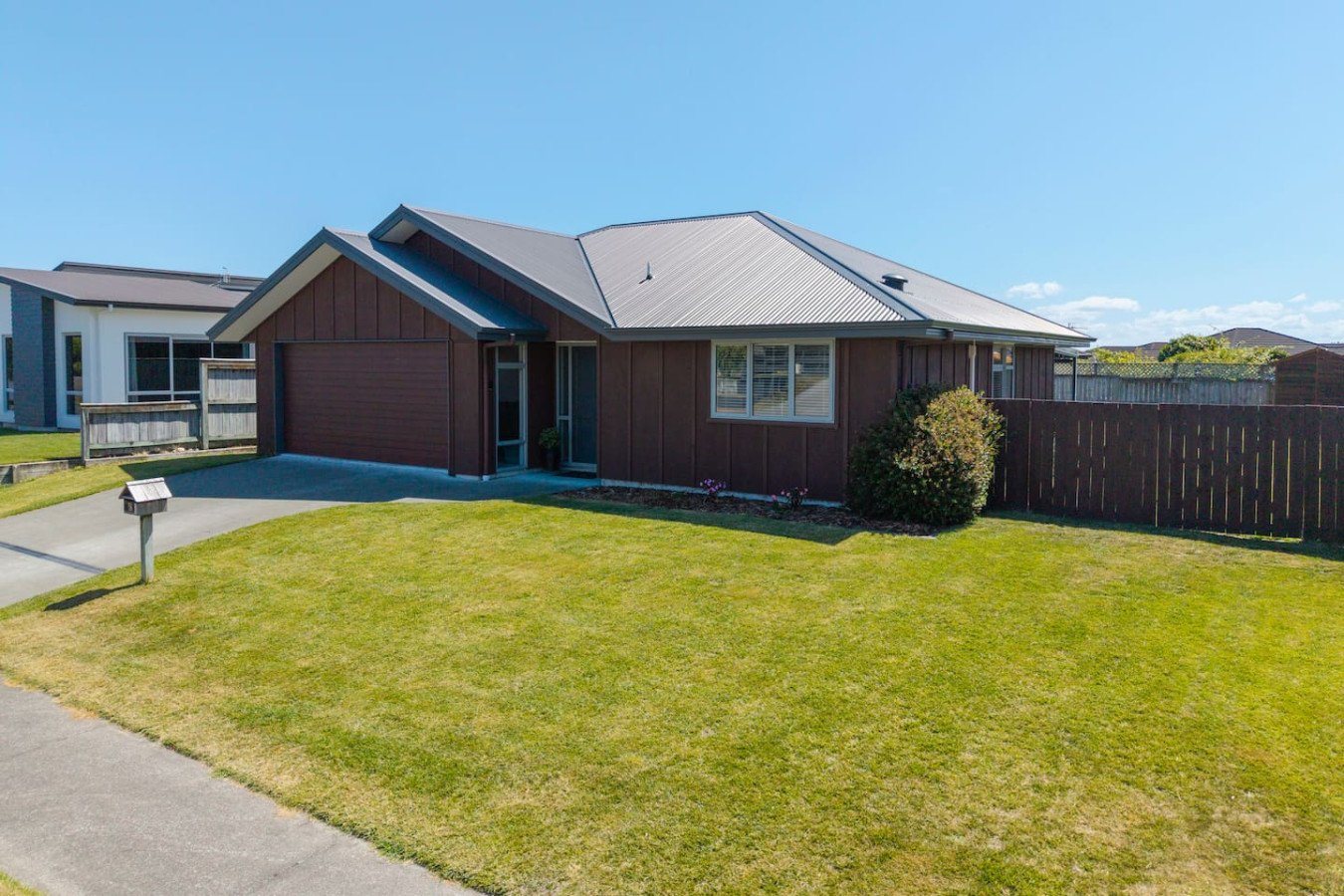 Front view of a single-story house with a brown exterior, metal roof, front yard with green grass, small bush, and a wooden fence on the right, overlooking a clear blue sky.