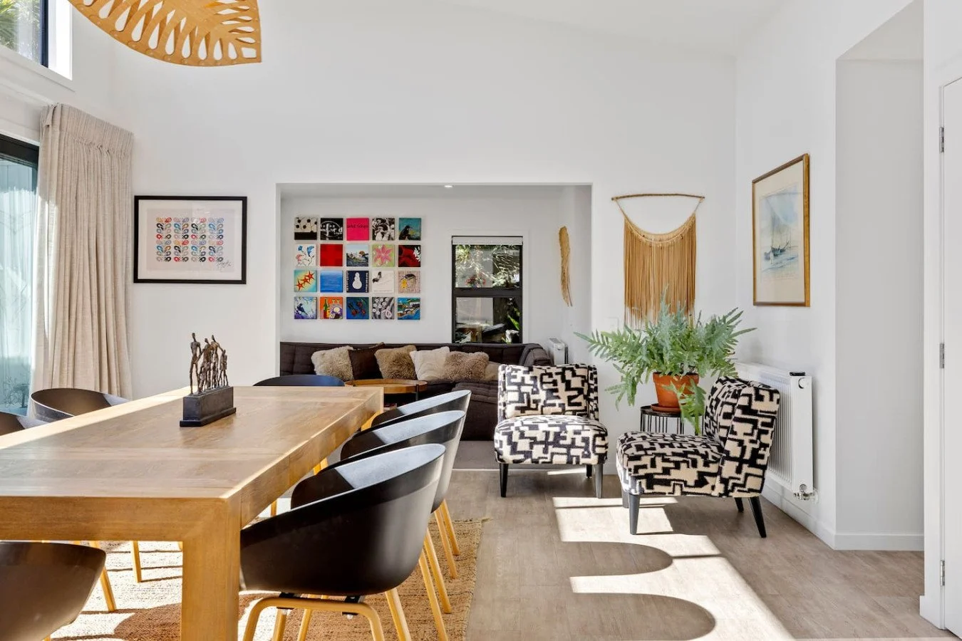 Bright, modern dining and living room with wooden table, black chairs, patterned armchairs, artwork on white walls, potted plant, and large windows with cream curtains.