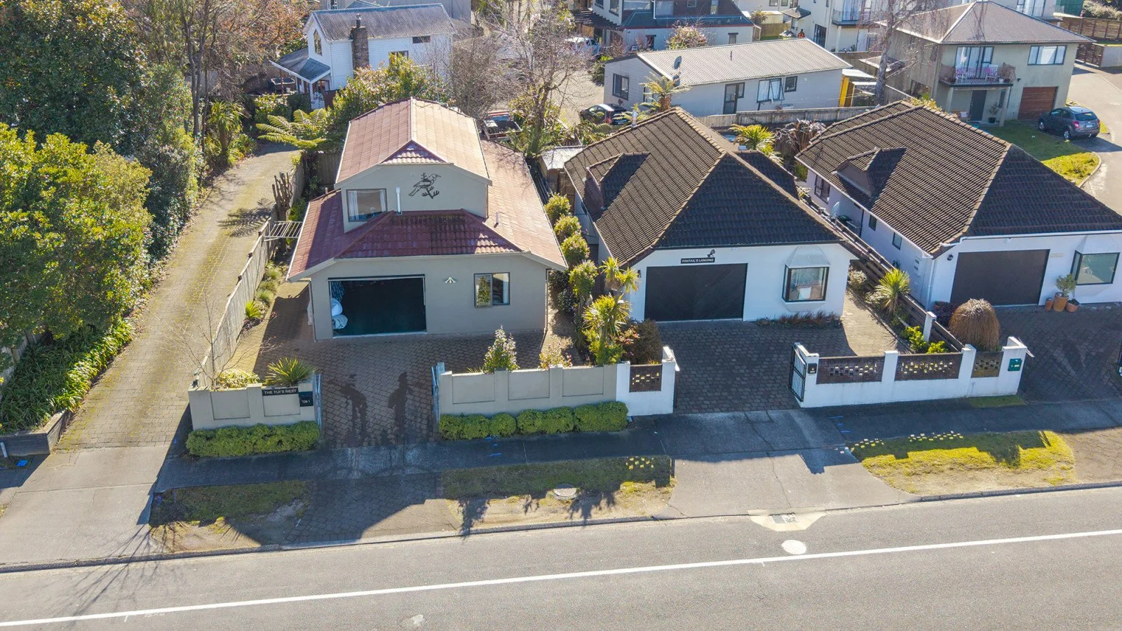Aerial view of three modern houses on a suburban street. The houses have driveways, fenced yards, and surrounding greenery, with trees and neighboring houses in the background.
