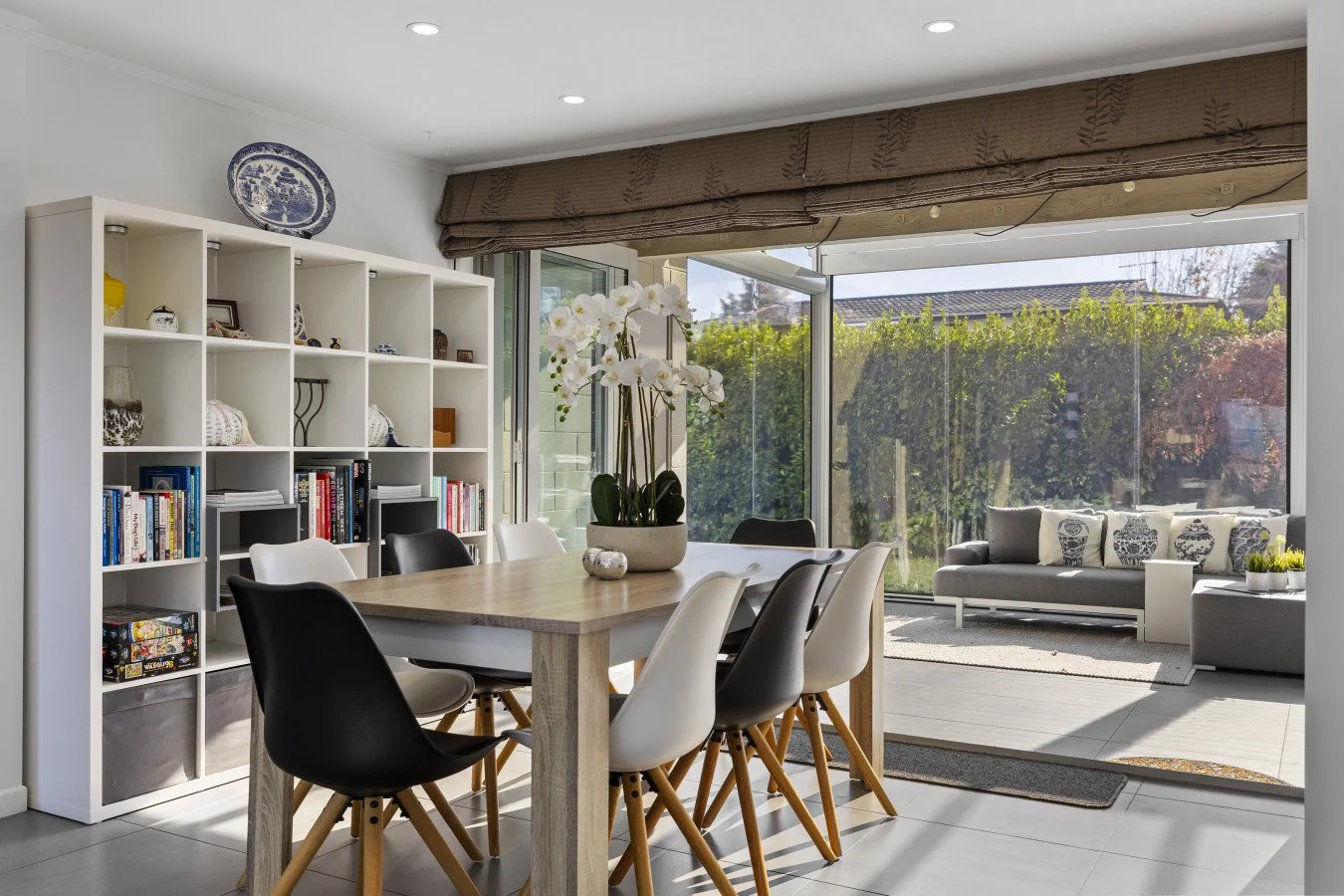 Bright dining area with a wooden table, black and white chairs, a white bookshelf with decorative items and books, a large orchid plant on the table, and a sunroom with a gray sofa and large windows showing greenery outside.