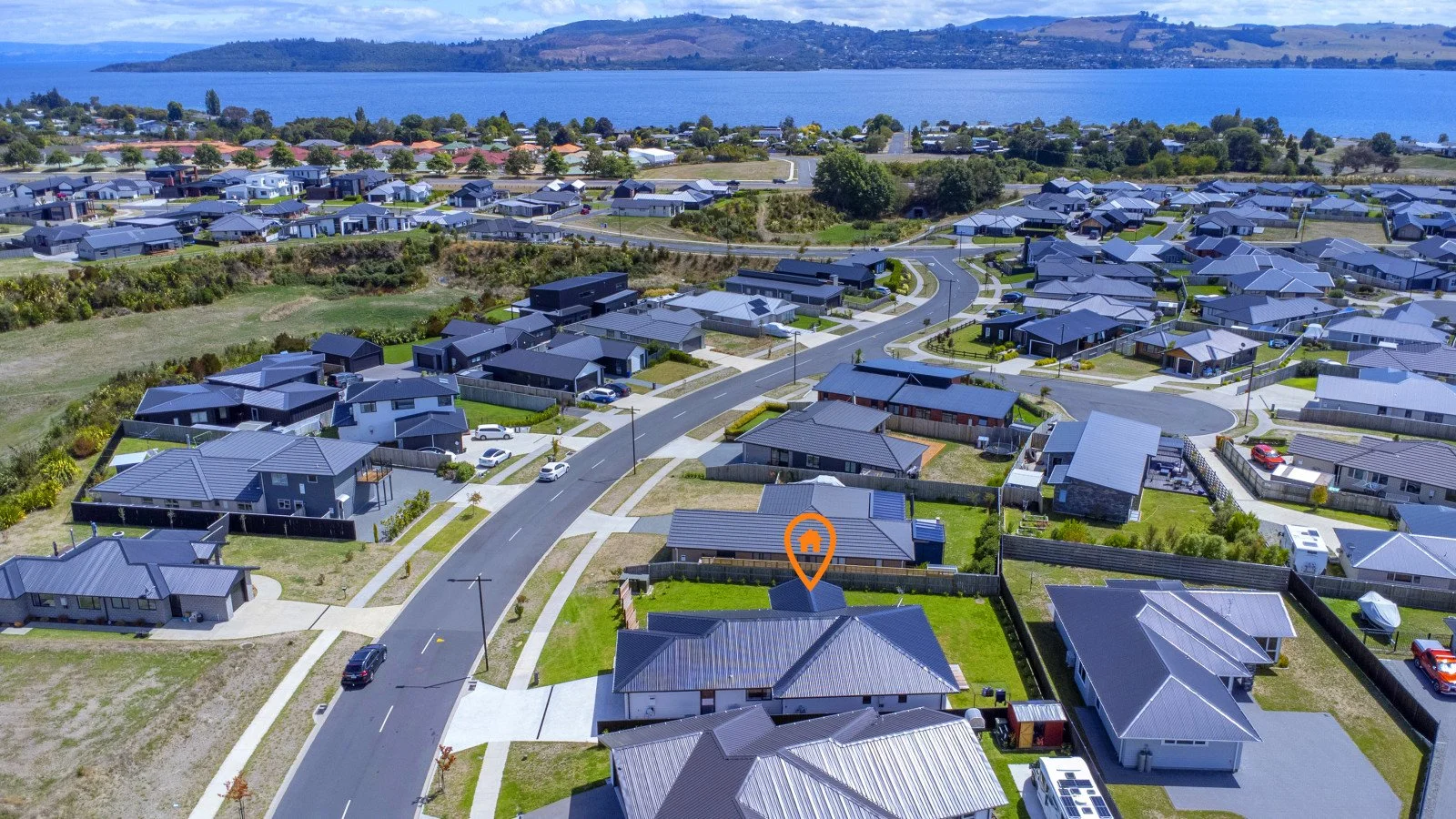 Aerial view of a suburban neighborhood with houses, streets, and yards by a large body of water, with hills in the background.
