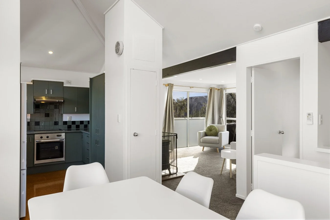 Interior of a modern apartment showing a kitchen with gray cabinets, a stove, backsplash, a dining area with a white table and chairs, and a living room with a white armchair, large windows, curtains, and a view of outdoors.