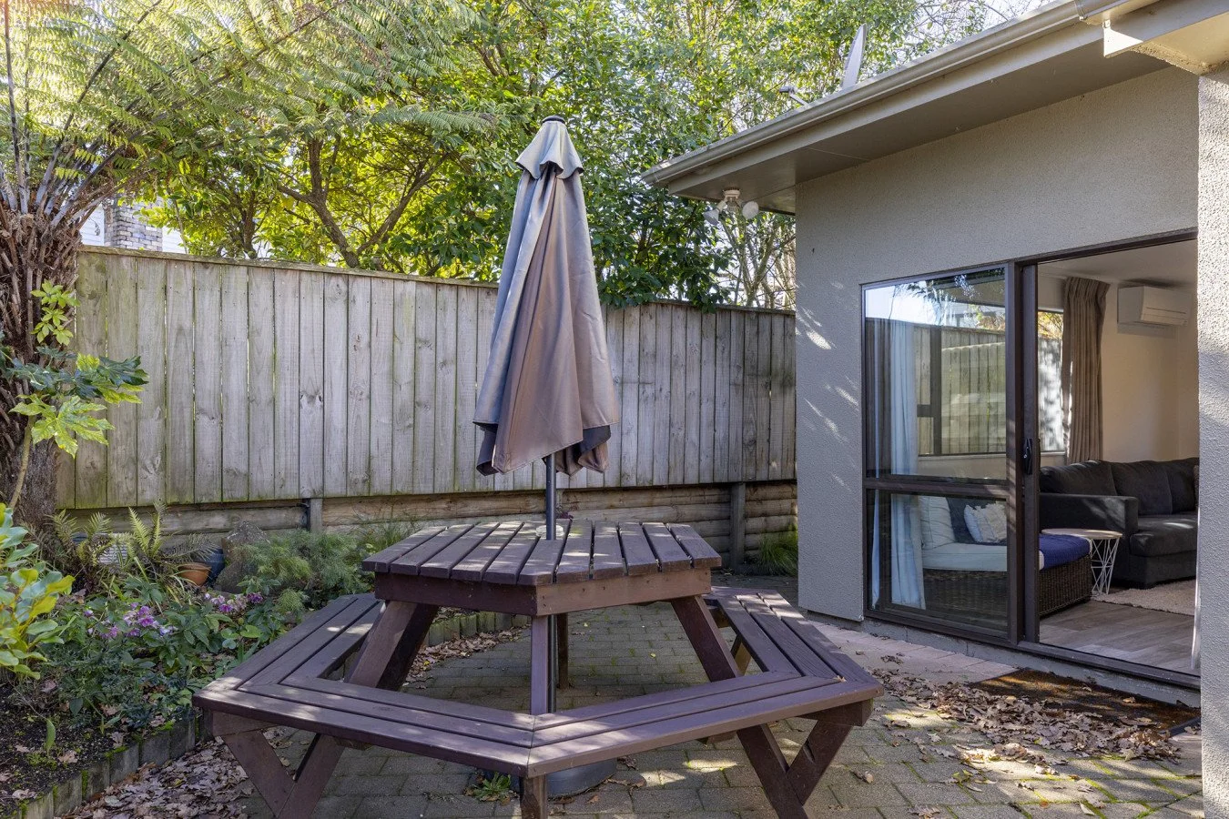 Backyard patio with wooden picnic table and closed umbrella, surrounded by a weathered wooden fence, greenery, and adjacent to a house with large sliding glass doors leading into a living room.