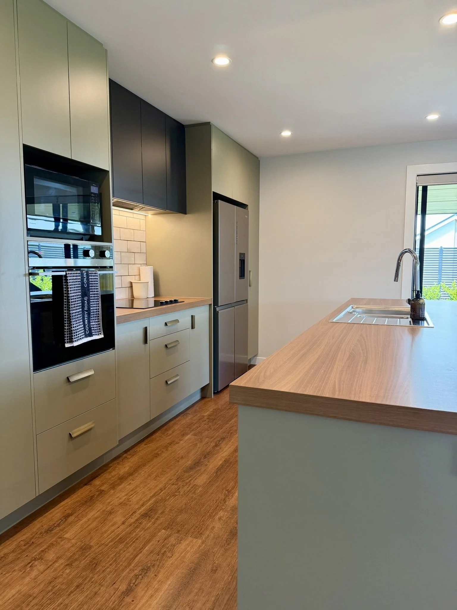 Modern kitchen with beige cabinets, black upper cabinets, stainless steel oven, microwave, and refrigerator, wooden countertop, and a window with blinds.