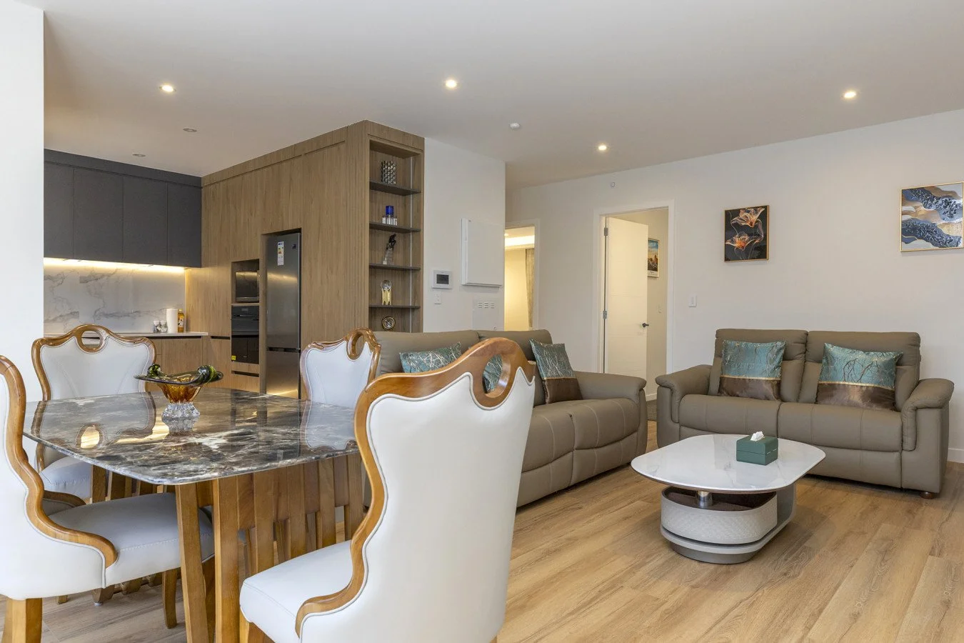 Modern living room with beige sofas, decorative pillows, a white marble coffee table, a dining table with a marble top and white upholstered chairs, and an open kitchen with dark cabinets, wooden accents, and marble backsplash.