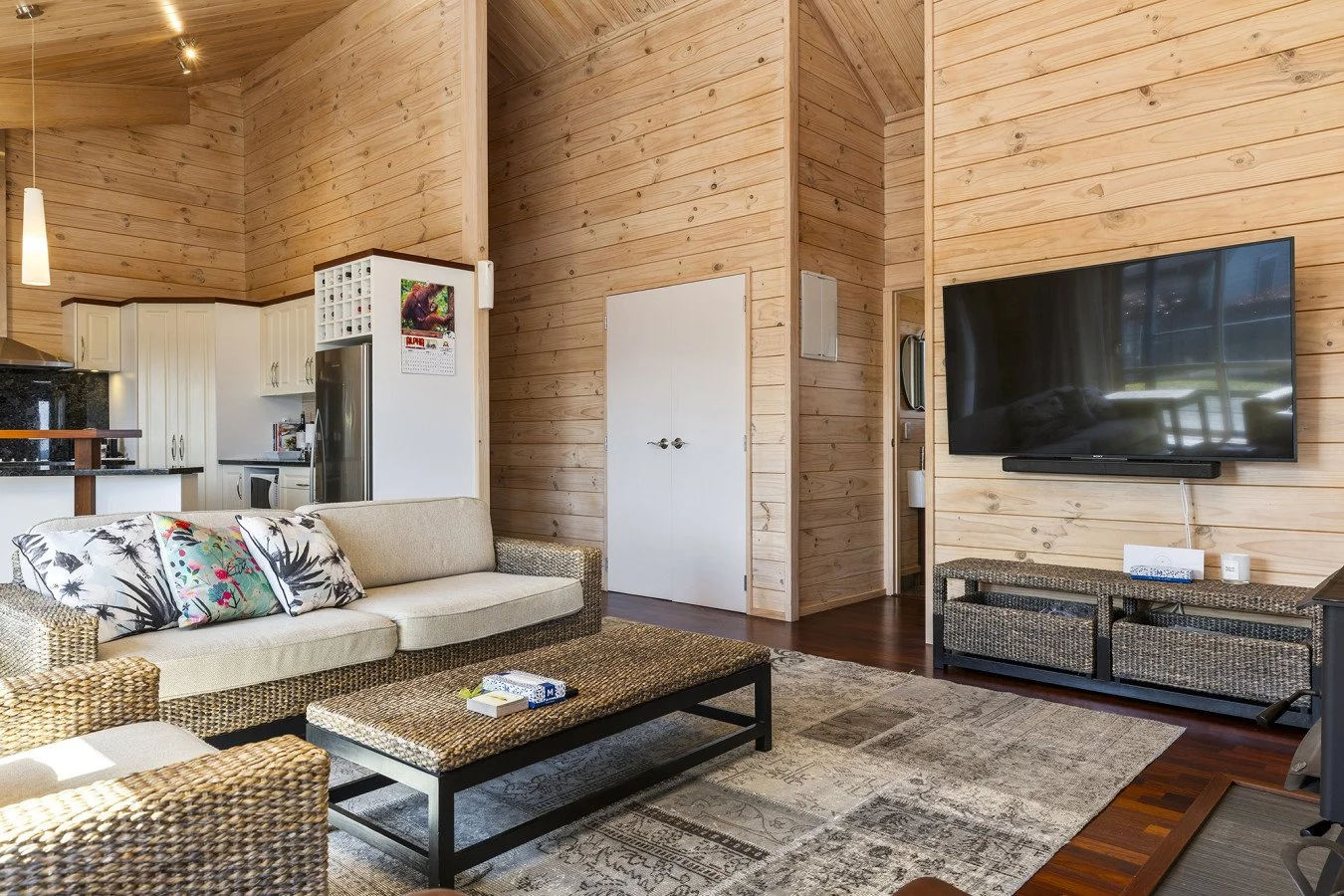 Living room with beige sofa, wicker armchair, wooden wall paneling, flat-screen TV on wall, woven TV stand, patterned area rug, coffee table, kitchen with white cabinets and stainless steel appliances in background.