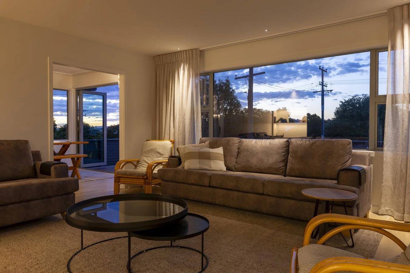 A cozy living room at dusk with large windows showing an outdoor view of trees and power lines, beige walls, plush sofas, wooden chairs, and a black coffee table.