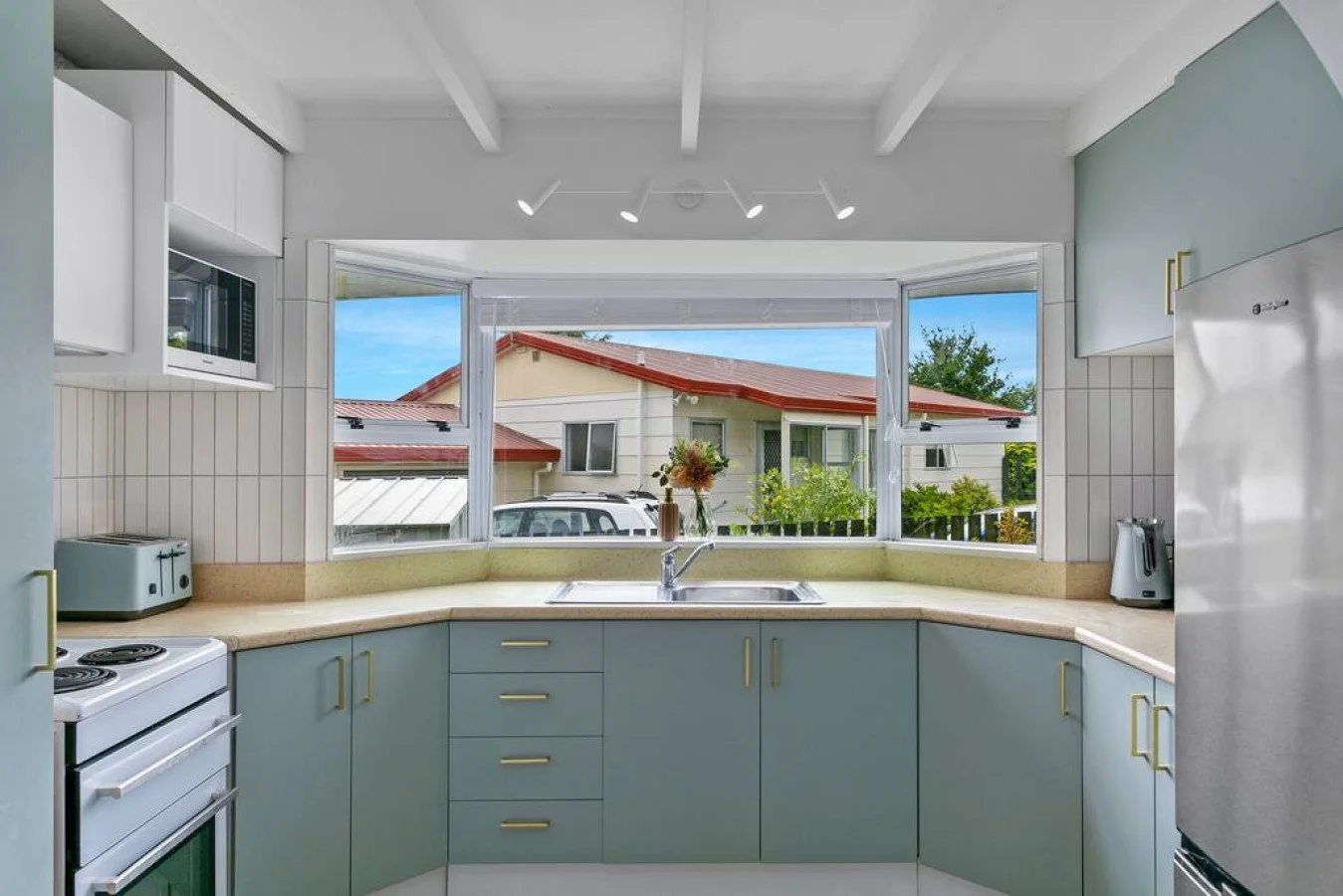 Kitchen with light blue cabinets, beige countertop, white appliance, and a double window overlooking a house with a red roof and trees.