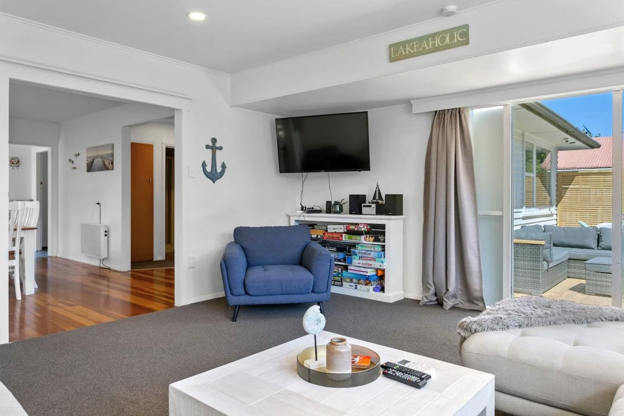 Living room with a blue armchair, a white coffee table with decorative items, a white sofa with a furry throw, a TV mounted on the wall, a bookcase with games, and sliding glass doors leading to an outdoor patio with seating.