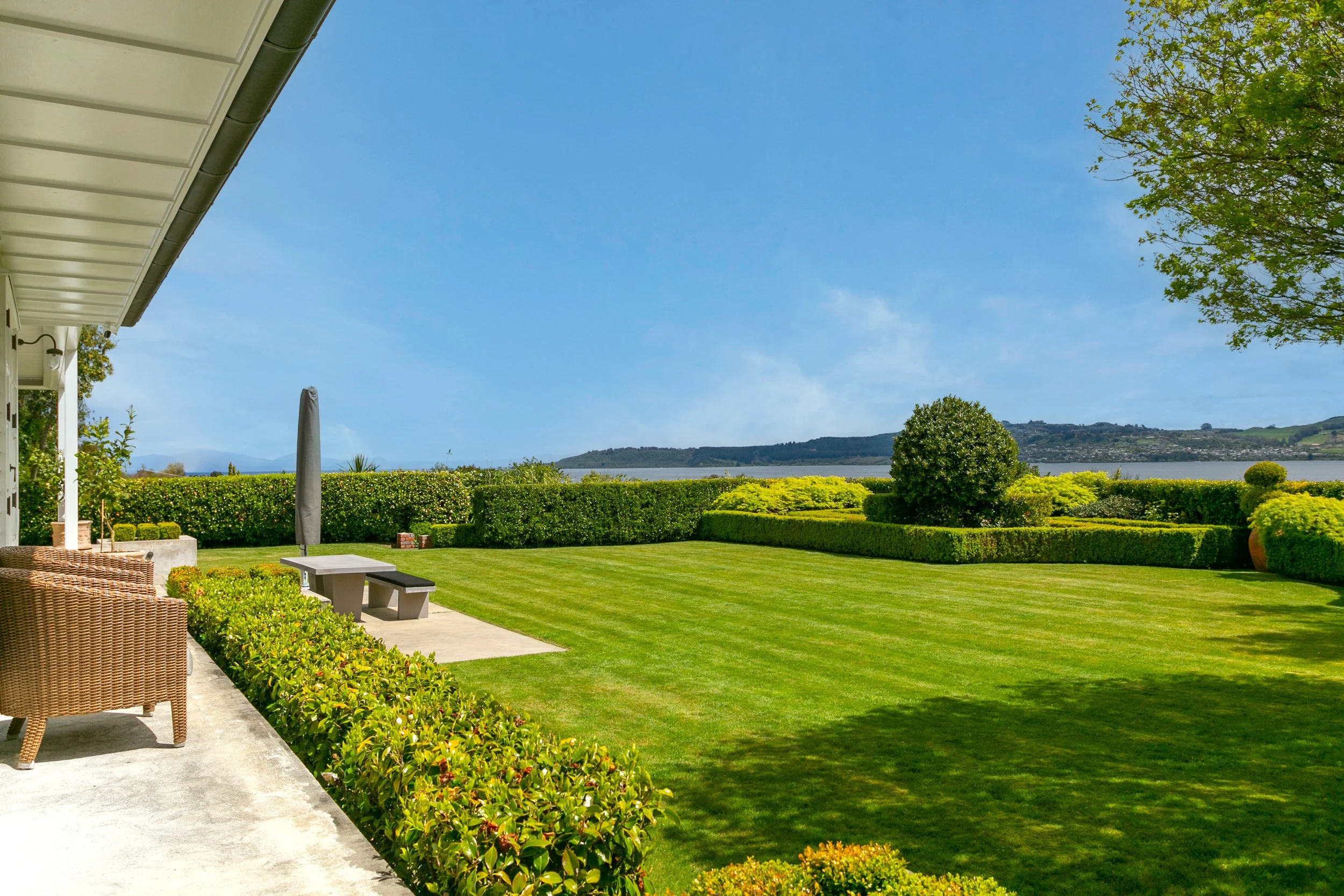 View of a lush, well-manicured lawn with shrubs and trees, overlooking a body of water and a hilly landscape in the distance, seen from a patio area with wicker chairs and a concrete path.