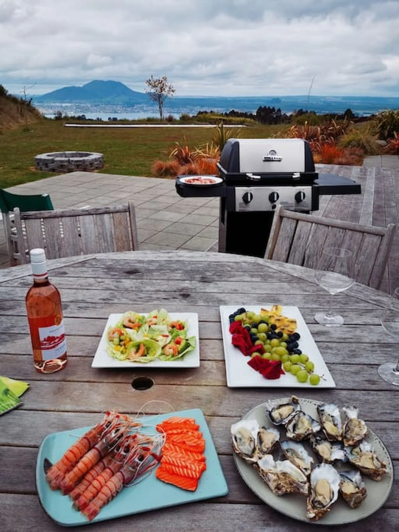 Outdoor dining setup with a wooden table holding seafood, fruit, salad, and drinks, with a barbecue grill and scenic landscape in the background.