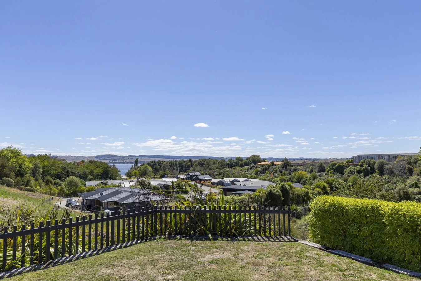 A landscape view of a neighborhood with houses, greenery, trees, and a body of water in the distance under a clear blue sky with scattered clouds.