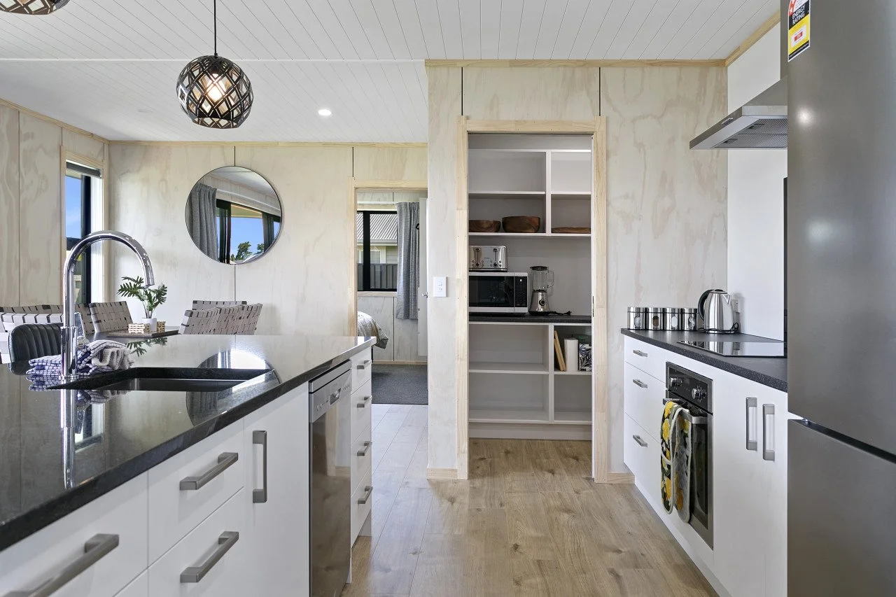 Modern kitchen with white cabinets, black countertops, a stainless steel refrigerator, and a small pantry with open shelves. There is a kitchen island with a sink and a black pendant light above. In the background, a dining area with a table and chai