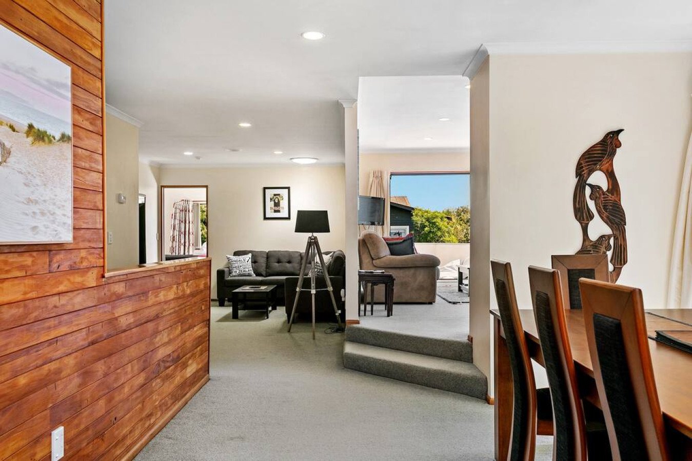 Living room with contemporary furniture, including a gray couch, a black armchair, and a beige armchair. There is a large window with a view of greenery outside, a wall-mounted artwork, and a wooden sculpture of three herons on the wall. A wooden din