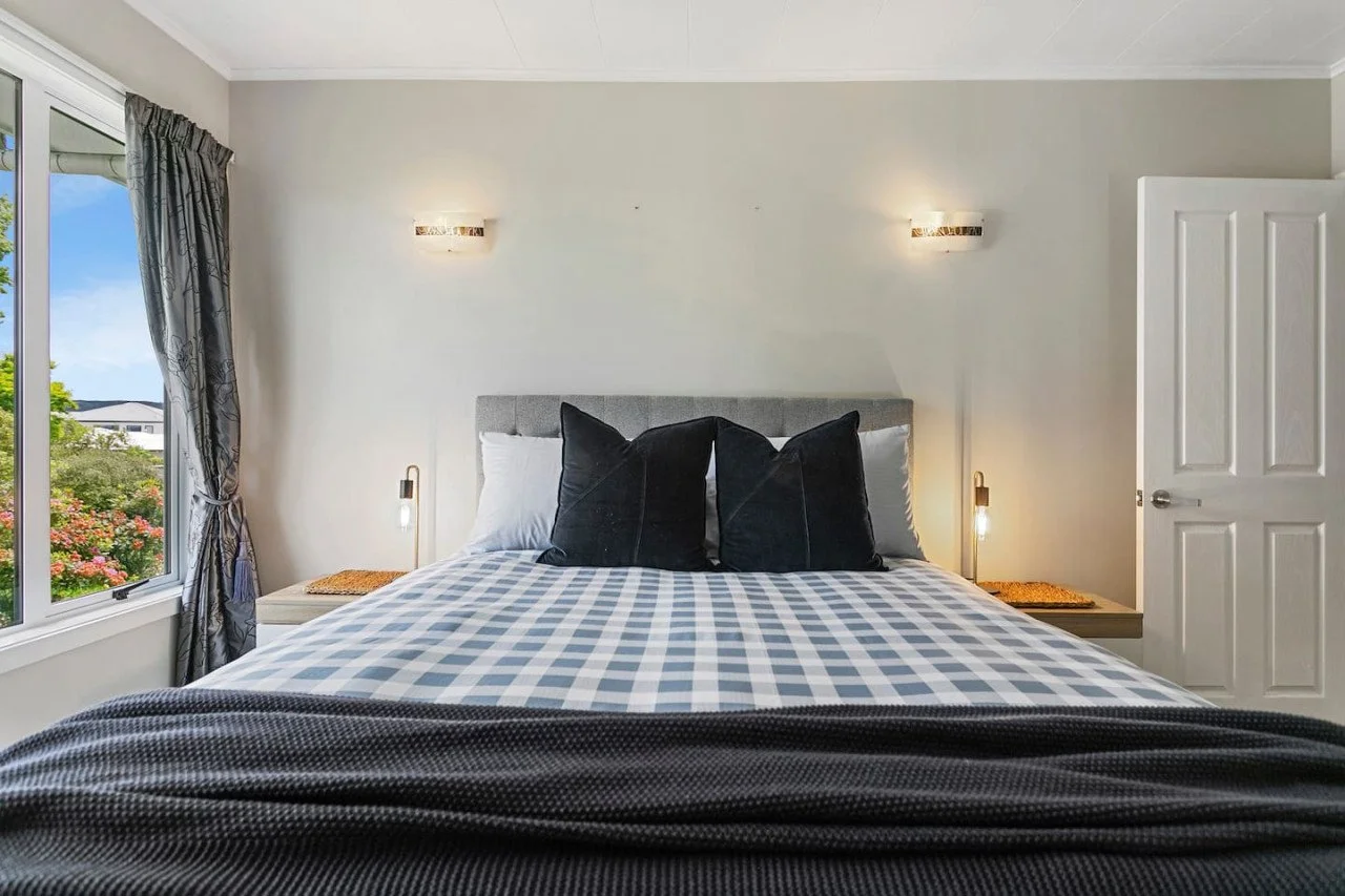 A neatly made bed with a draped black blanket, white sheets, and three black pillows, placed in front of a light-colored wall with two wall-mounted lights. A window with patterned curtains shows a view of trees and a cloudy sky outside.
