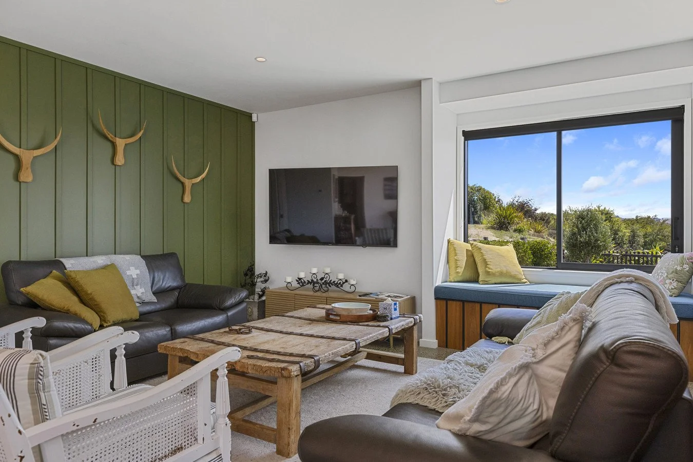 Living room with a green accent wall decorated with three wooden antler mounts, a black leather sofa with yellow and gray pillows, a rustic wooden coffee table, a wall-mounted TV, a white chair with pillows, and a large window with a view of trees an