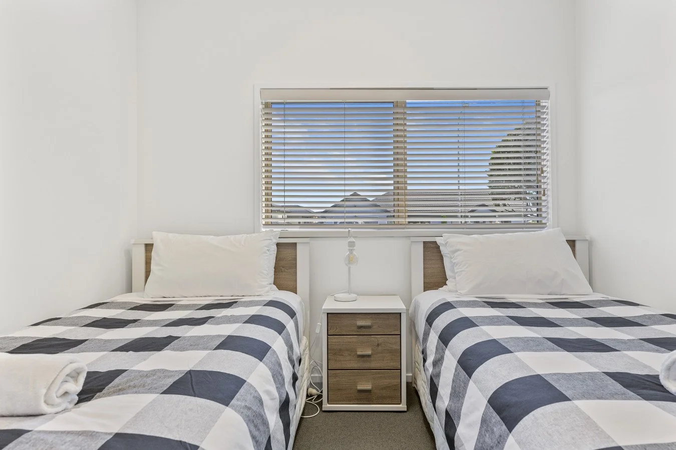 Two twin beds with black and white checkered bedding and white pillows, a nightstand with drawers and a lamp between them, in a room with a window with blinds showing rooftops and trees outside.