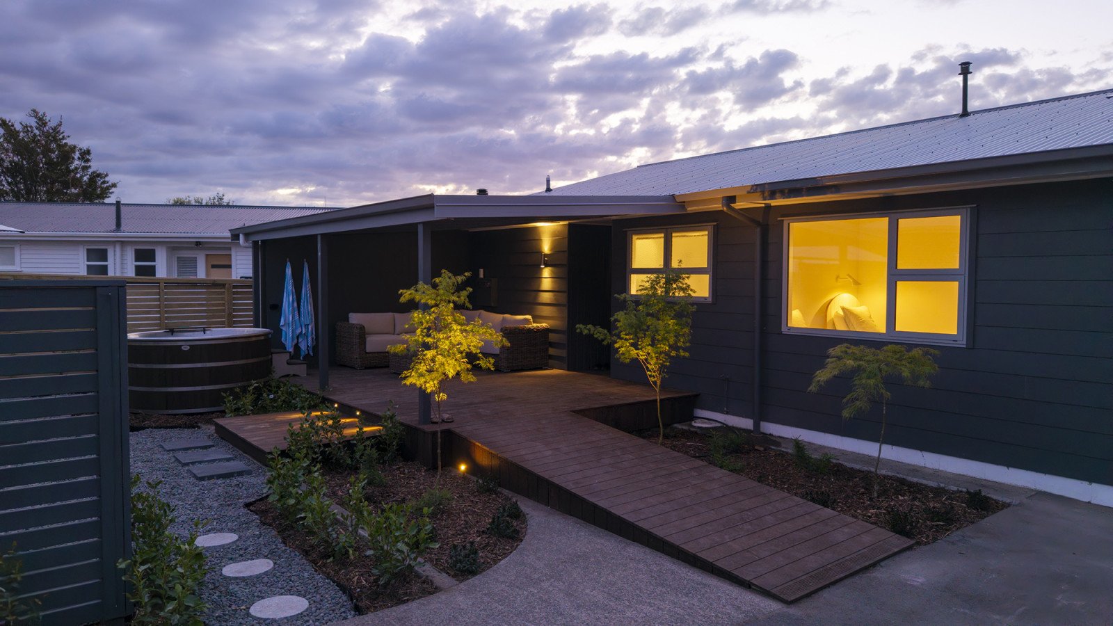 A backyard patio area at dusk with outdoor furniture, a hot tub, a wooden walkway, small trees, and glowing interior windows of a house.