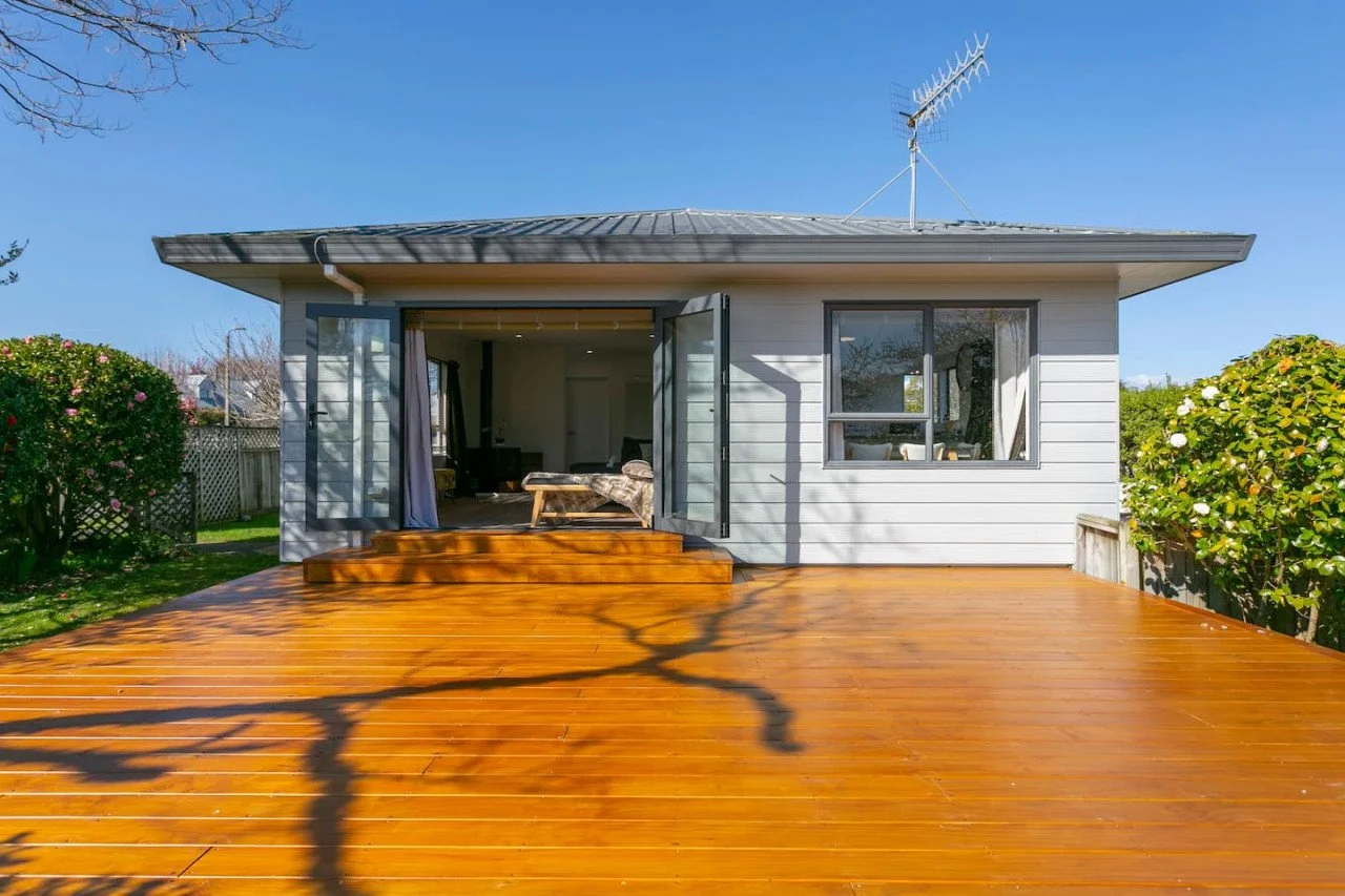 Backyard of a house with a wooden deck, open sliding glass doors, and a window, featuring bushes on either side and a TV antenna on the roof under a clear blue sky.