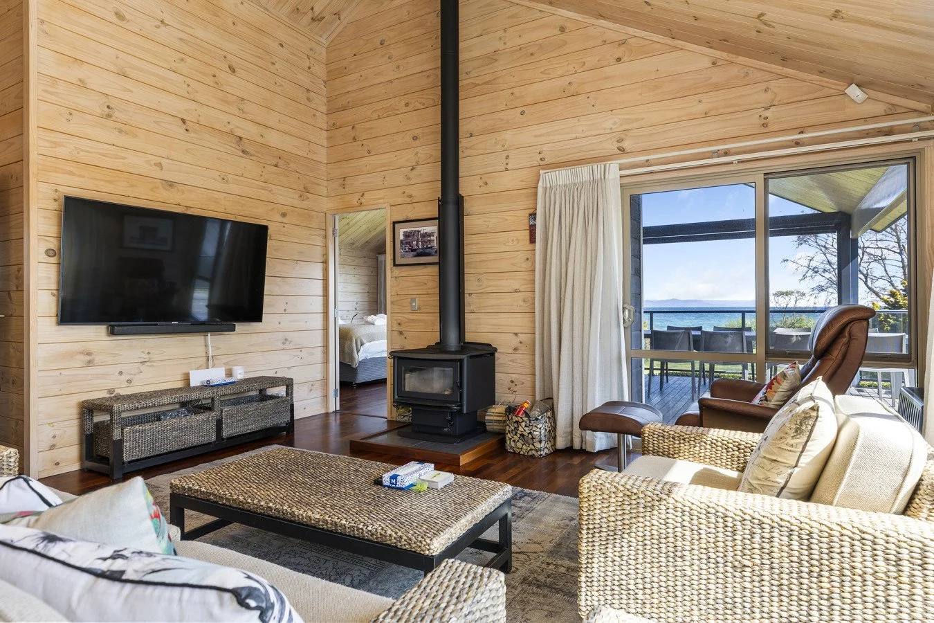 Living room with wood-paneled walls, a flat-screen TV on the wall, a wood stove with a chimney, and a sliding glass door leading to a balcony view of water and trees.