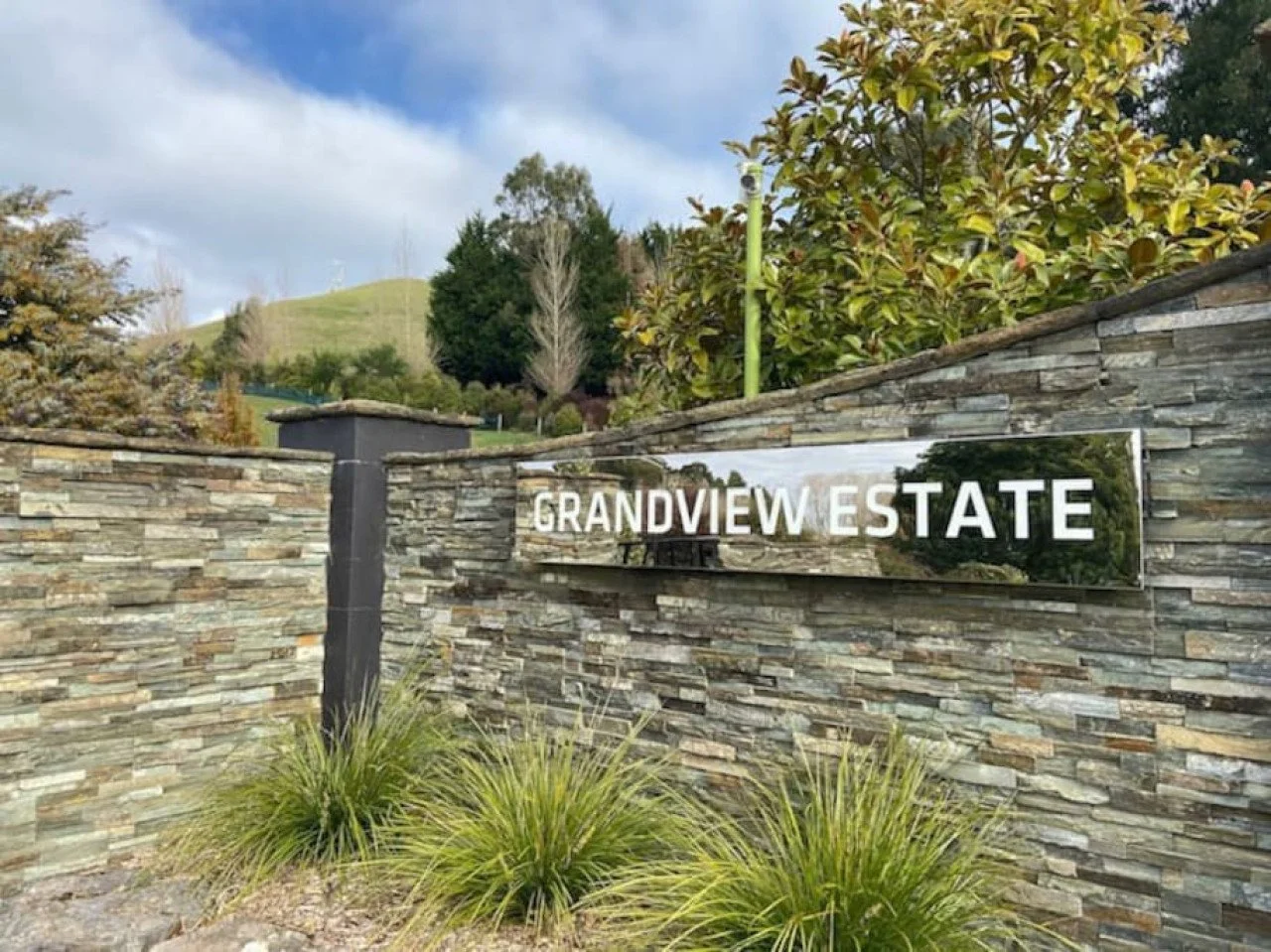 A stone wall with a reflective silver sign that reads 'Grandview Estate' surrounded by greenery and trees.