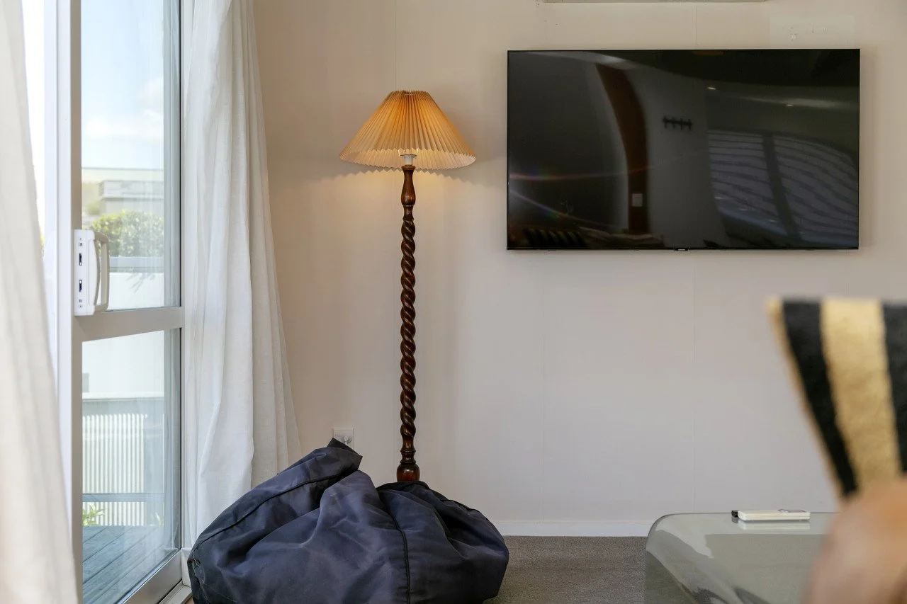 Living room with a sliding glass door, beige curtains, a tall decorative floor lamp, a large flat-screen TV, a black bag on the floor, and a table with a remote control.