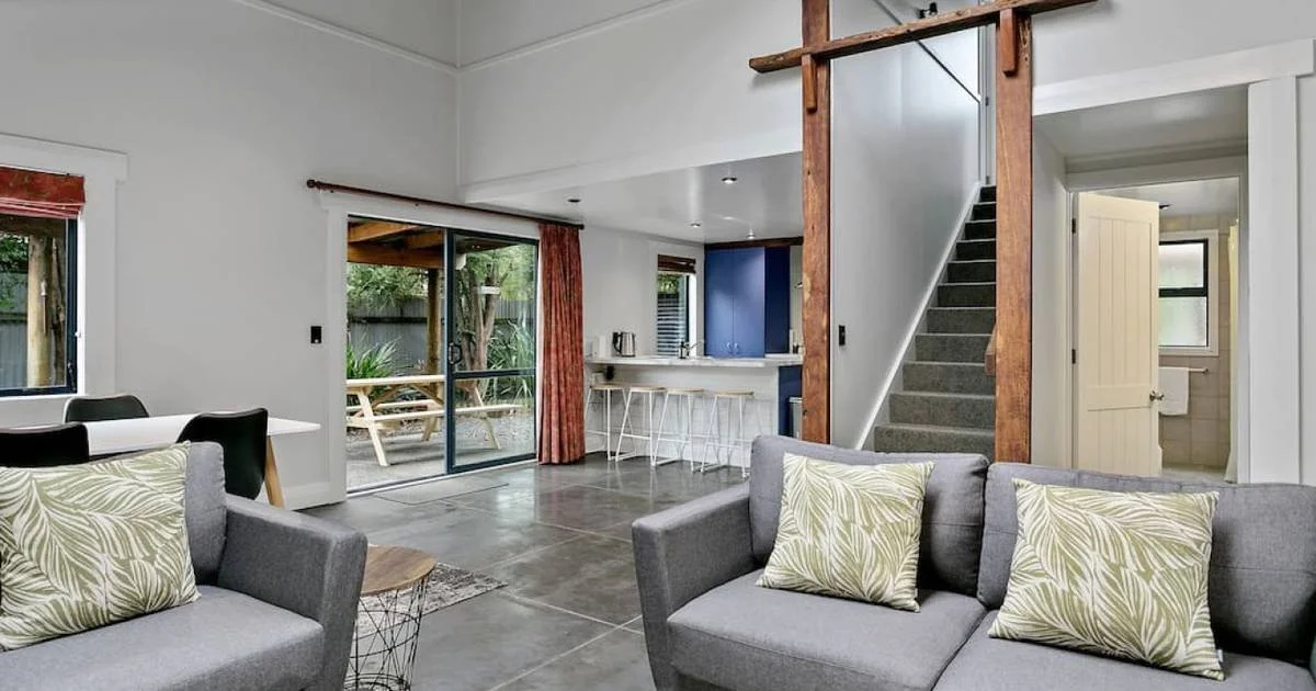 Living room with gray sofas, patterned pillows, a dining table with black chairs, and a deck outside visible through sliding glass doors. There is a staircase with wooden framing, a small kitchenette, and a bathroom.
