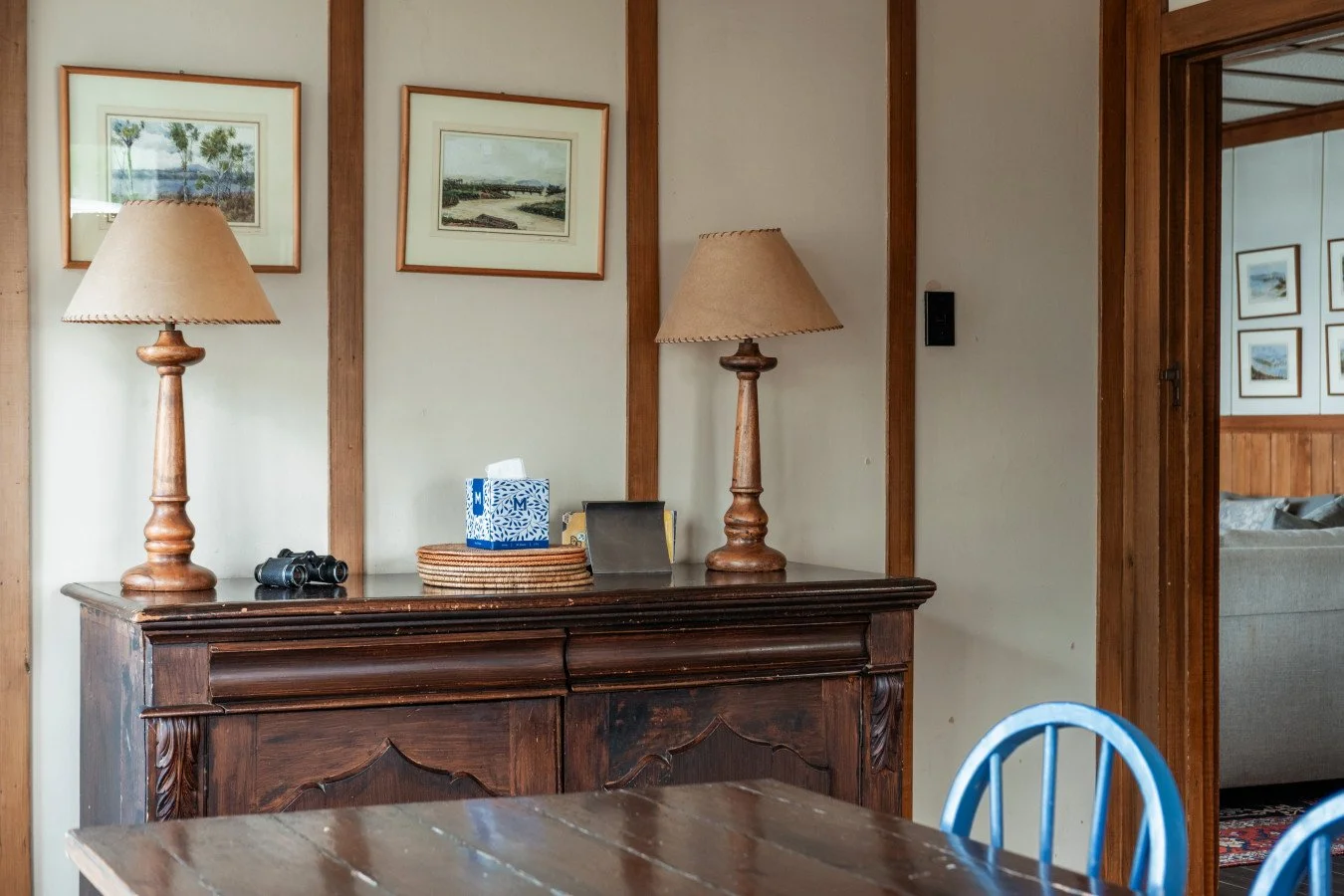 A wooden sideboard with two table lamps, a tissue box, binoculars, and a framed photo. Framed landscape paintings hang on the wall above. A doorway leads to a room with a gray couch and framed pictures on the wall.