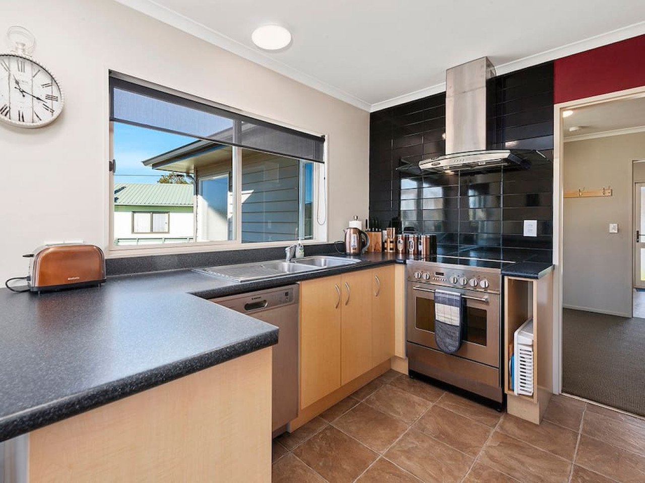 Modern kitchen with black countertops, light wooden cabinets, and a stainless steel stove with hood. There is a window above the sink and a clock on the wall.