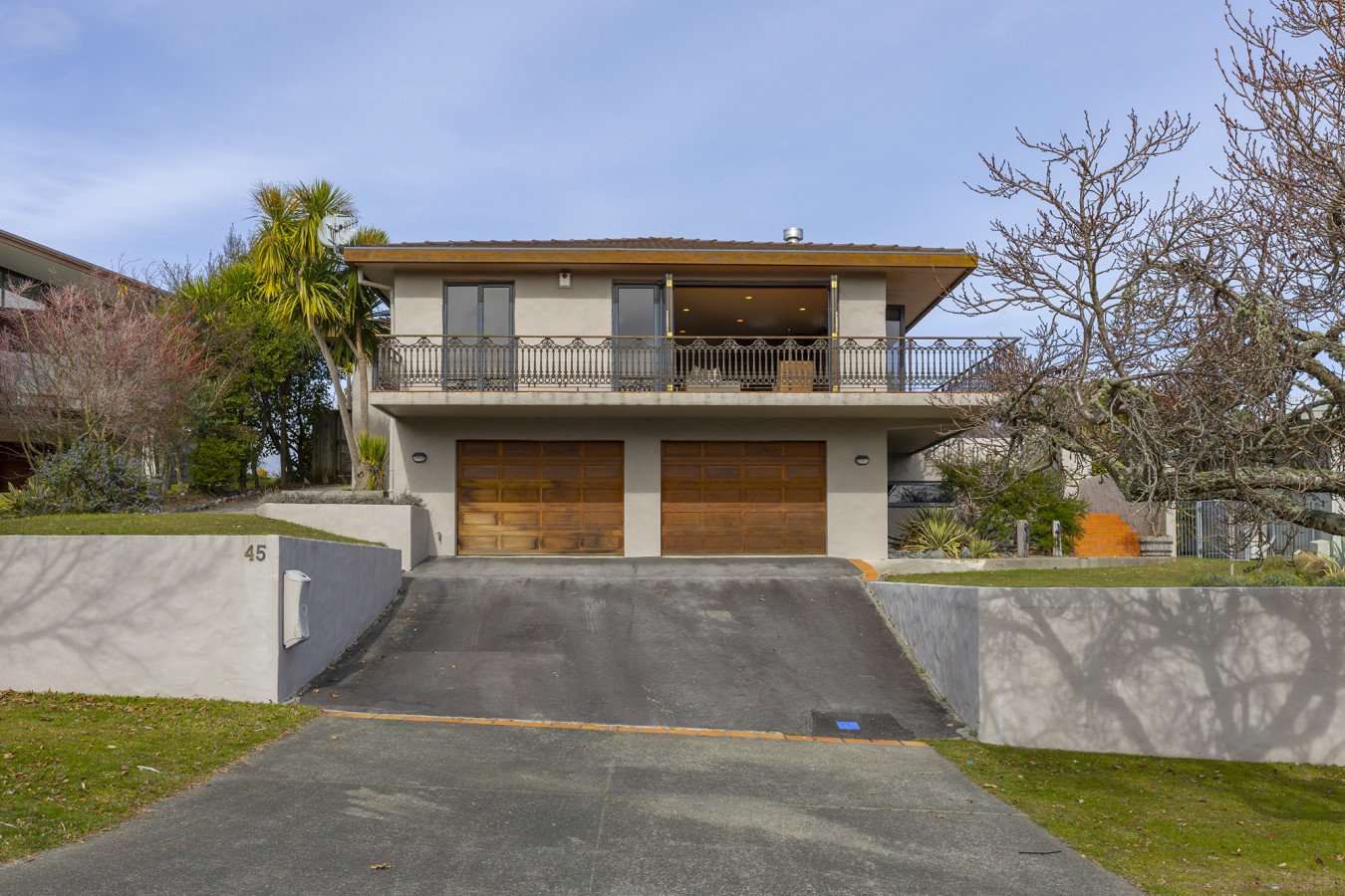 Front view of a modern two-story house with garages, a driveway, and a balcony, surrounded by trees and landscaping.