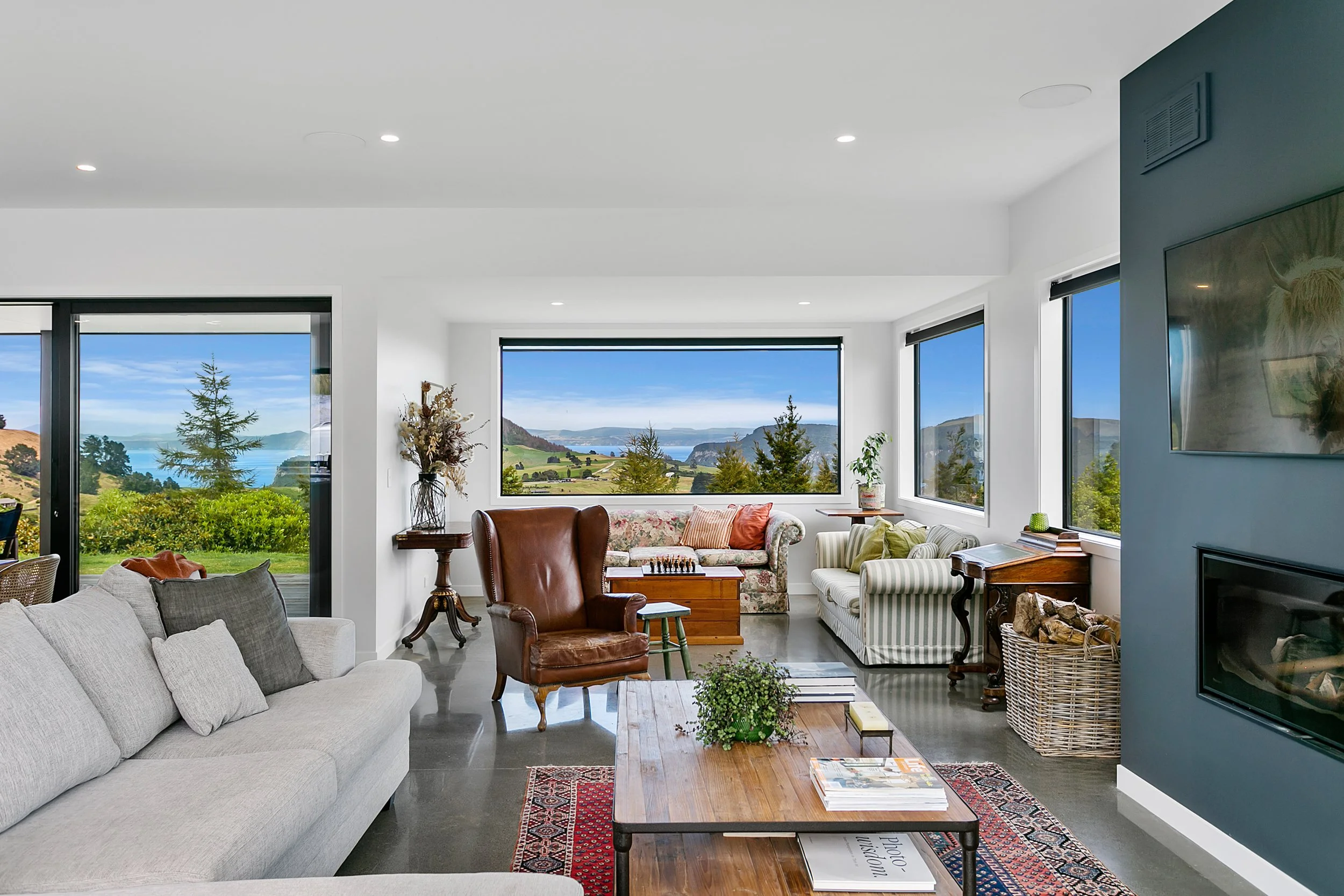 Living room with large windows showing scenic landscape, featuring sofas, armchair, coffee table, and decorative plants.